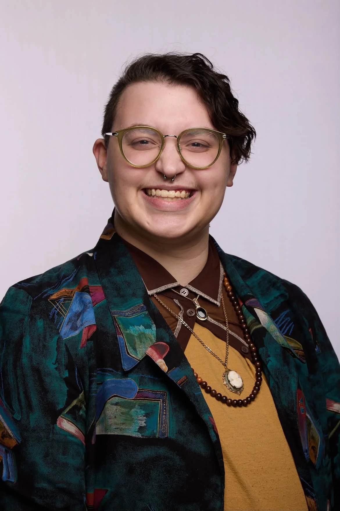 A person with short wavy hair, glasses, and multiple necklaces smiling. They are wearing a colorful patterned jacket over a brown shirt and a mustard yellow top, with a septum piercing against a plain, light background.