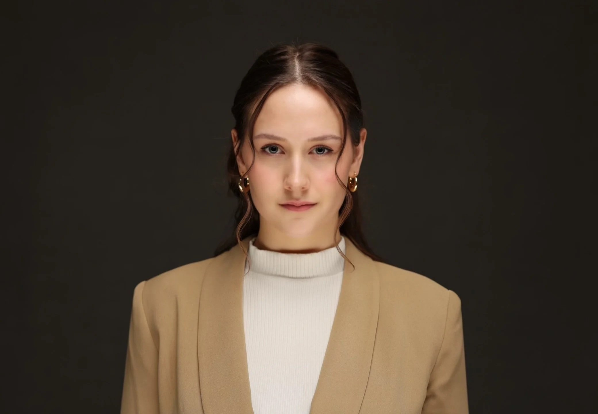 A young woman with brown hair styled in loose waves, wearing a beige blazer over a white turtleneck, and gold hoop earrings, looking directly at the camera against a dark background.