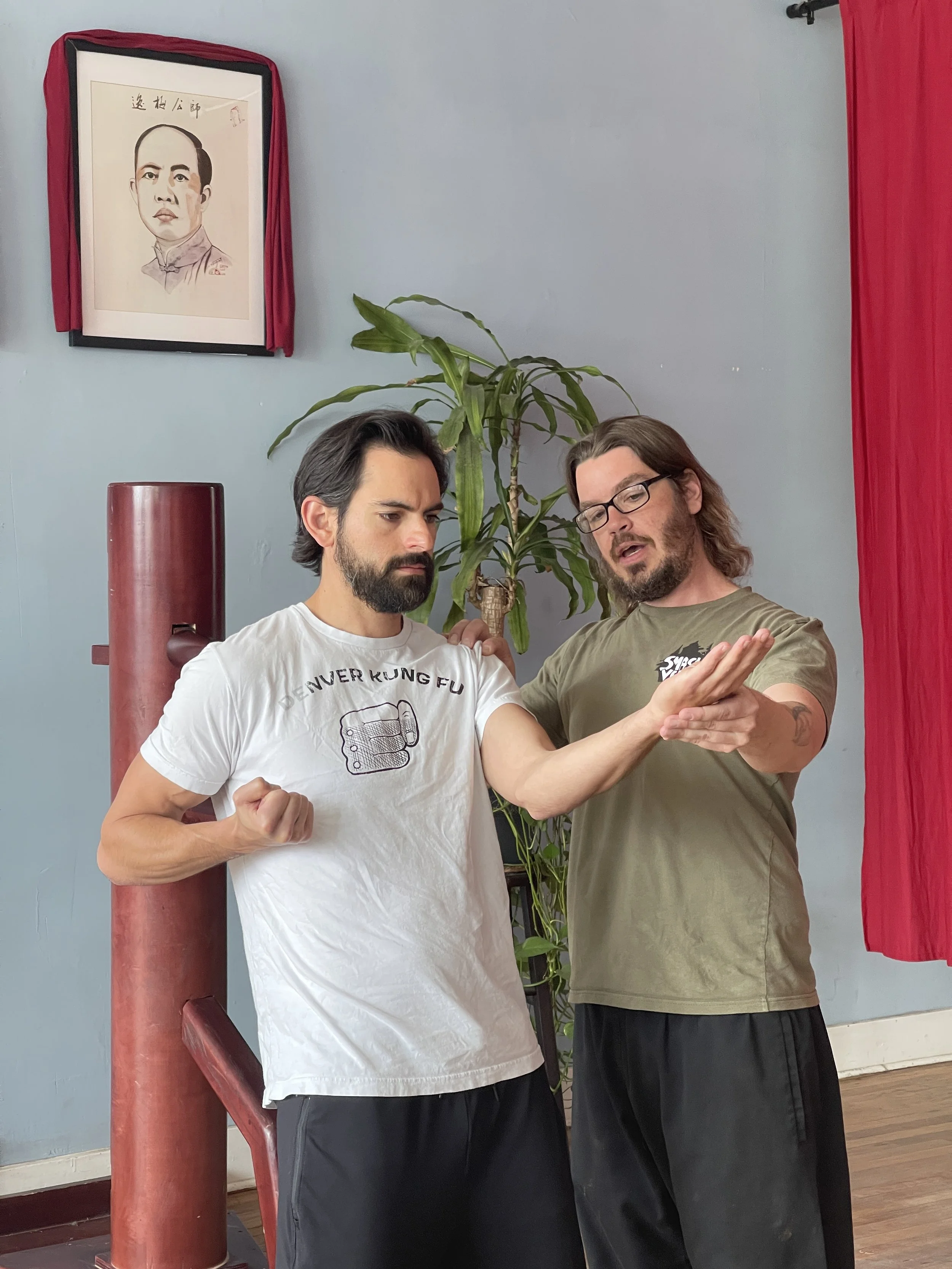 Two men practicing martial arts together indoors, standing near a red punching bag, with a framed painting of a man in traditional Chinese attire with Chinese characters above him on the wall behind them, next to a potted plant.