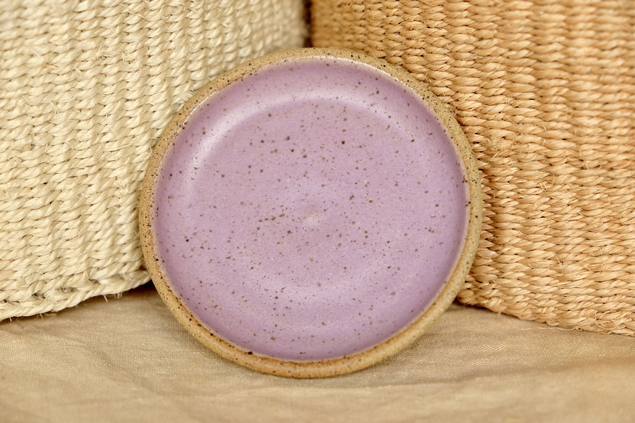 Hand-thrown ceramic ring dish glazed with lilac glaze, resting against warm tablecloth background.