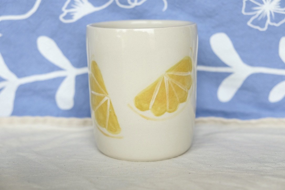 A hand-thrown porcelain tumbler, painted with lemon slices, sits on a cream colored tablecloth.