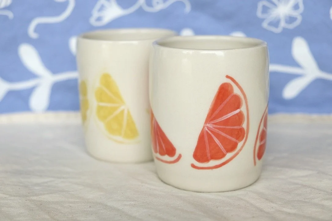 Two hand-thrown porcelain tumblers sit side by side, one hand-painted with lemon slices and the other with grapefruit slices, on a cream colored linen cloth.