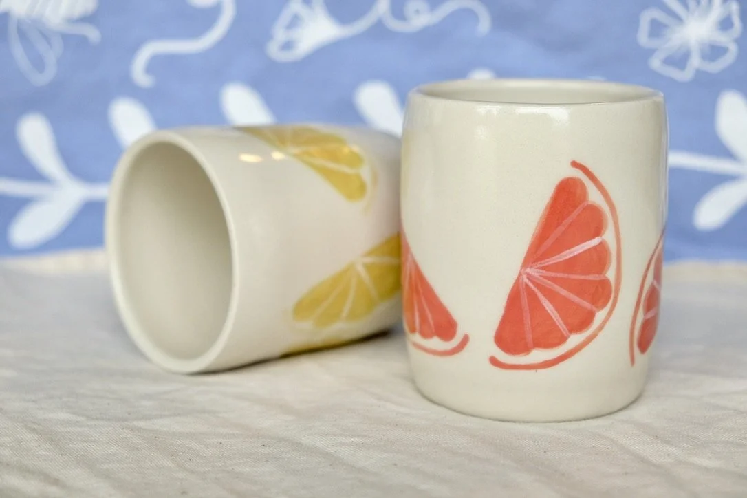 Two hand-thrown porcelain tumblers, one on its side and painted with lemon slices, and the other upright and painted with grapefruit slices, sit on a cream colored tablecloth.