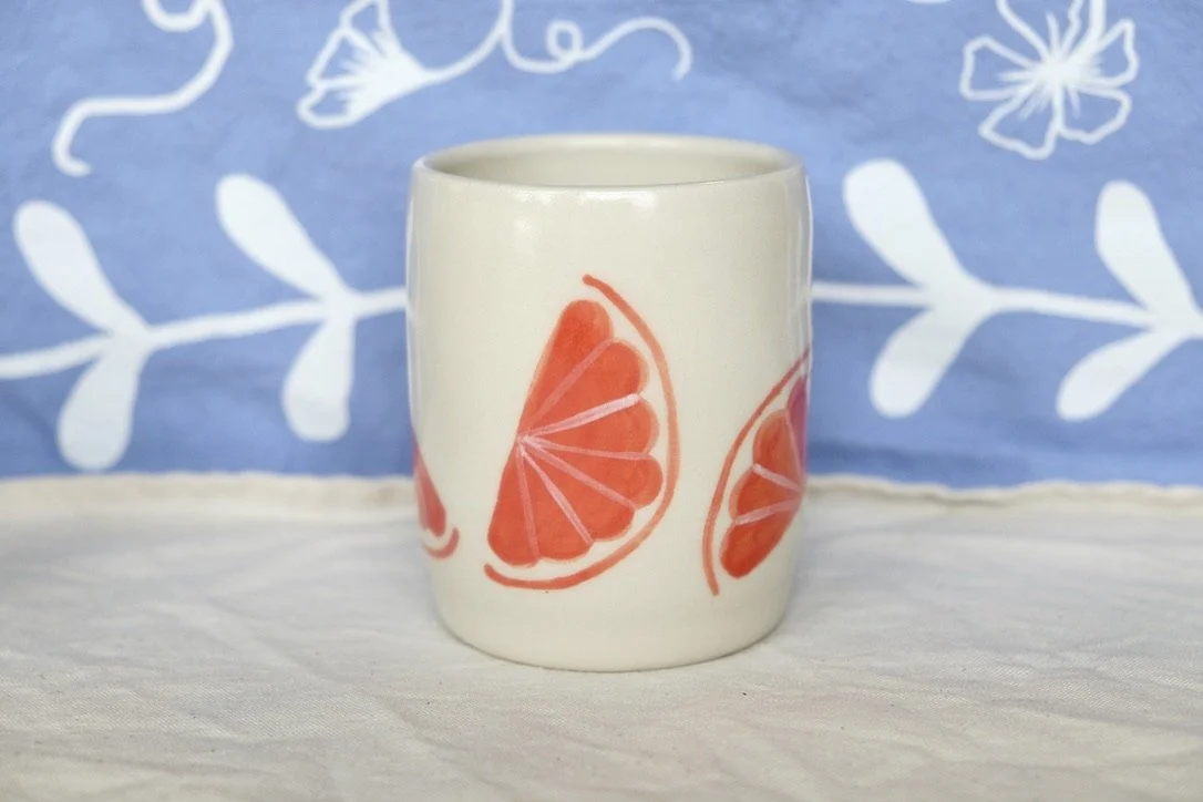 A hand-thrown porcelain tumbler, painted with grapefruit slices, sits on a cream colored tablecloth.
