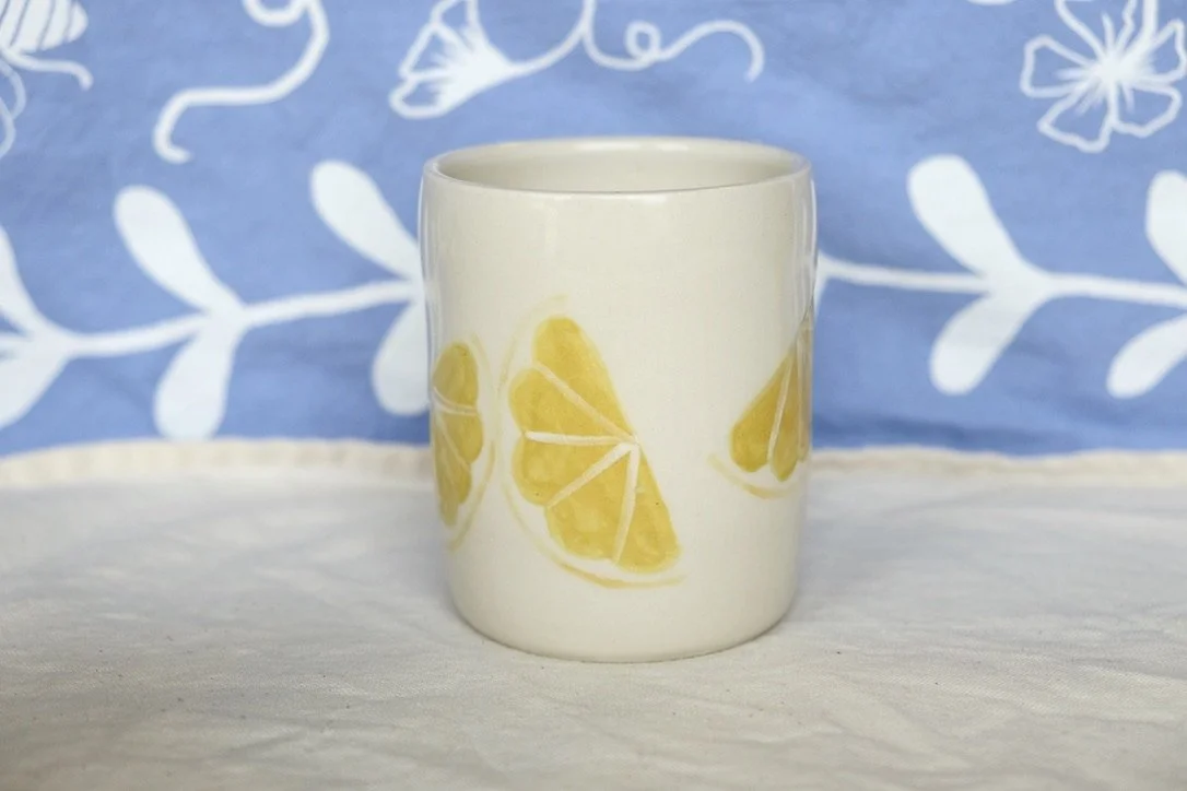 A hand-thrown porcelain tumbler, painted with lemon slices, sits on a cream colored tablecloth.