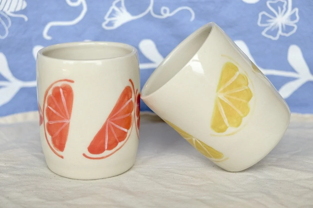 Two hand-thrown porcelain tumblers, one leaned against the other and painted with lemon slices, and the other upright and painted with grapefruit slices, sit on a cream colored tablecloth.