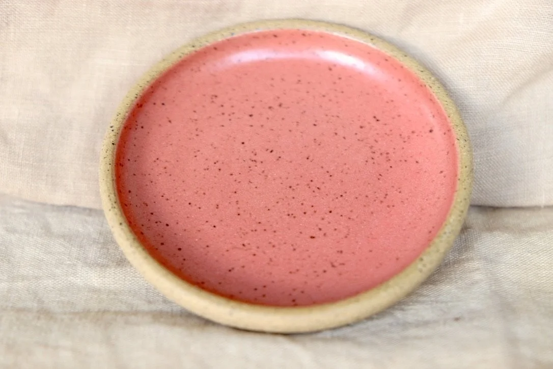 Hand-thrown ceramic ring dish glazed with coral glaze, resting against warm tablecloth background.