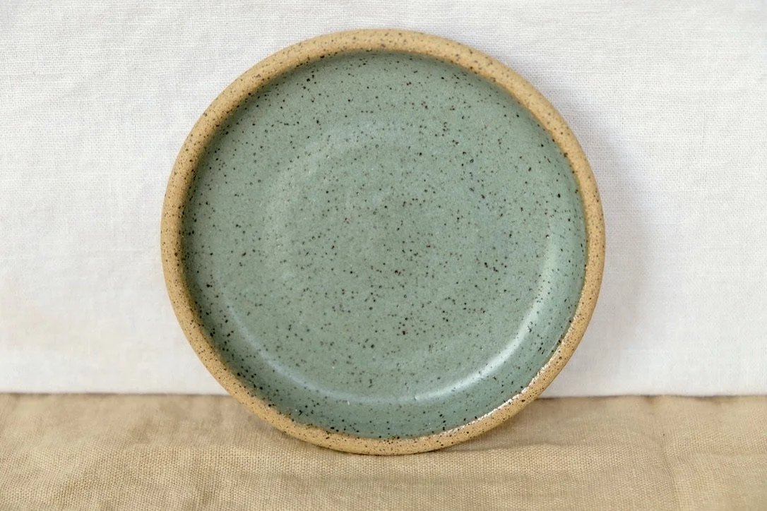 Hand-thrown ceramic ring dish glazed with avocado glaze, resting against warm tablecloth background.