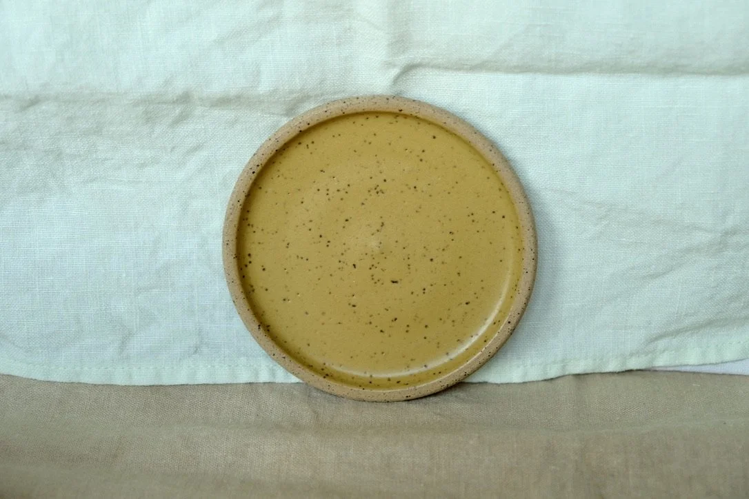 Hand-thrown ceramic ring dish glazed with mustard glaze, resting against warm tablecloth background.