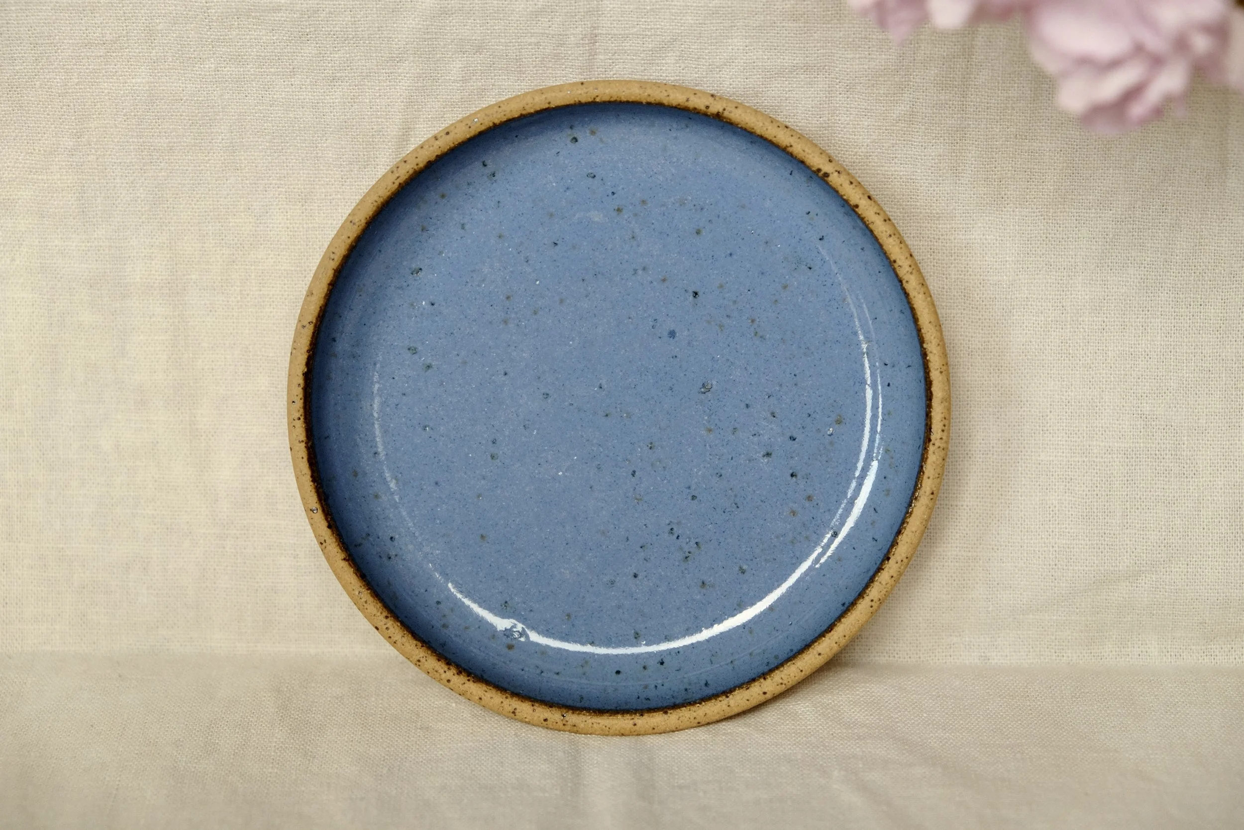 Hand-thrown ceramic ring dish glazed with cornflower blue glaze, resting against warm tablecloth background.