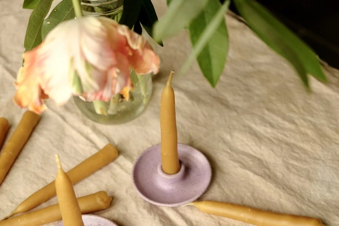 Hand-dipped beeswax taper candles and lilac colored ceramic candle holders lay on a tan table cloth.