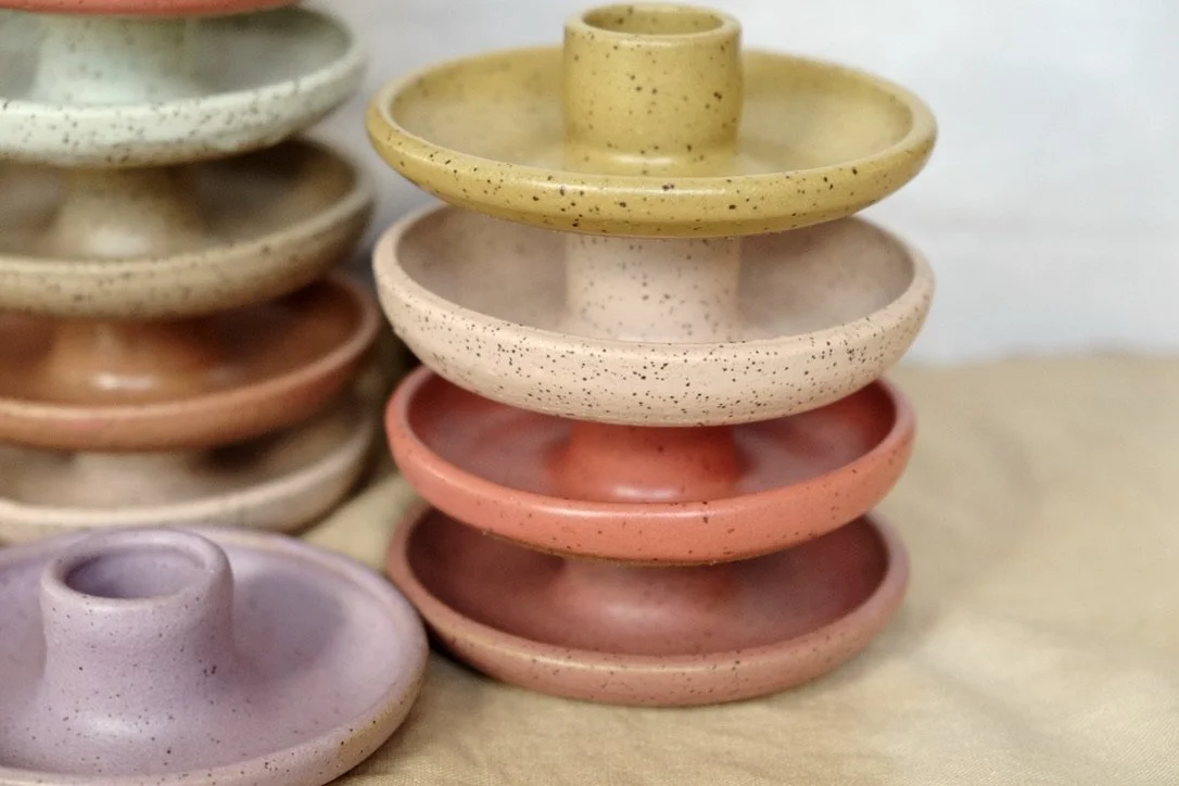 Four hand-thrown circular ceramic candle holders are stacked on top of each other, in mustard yellow, pink, coral, and red.