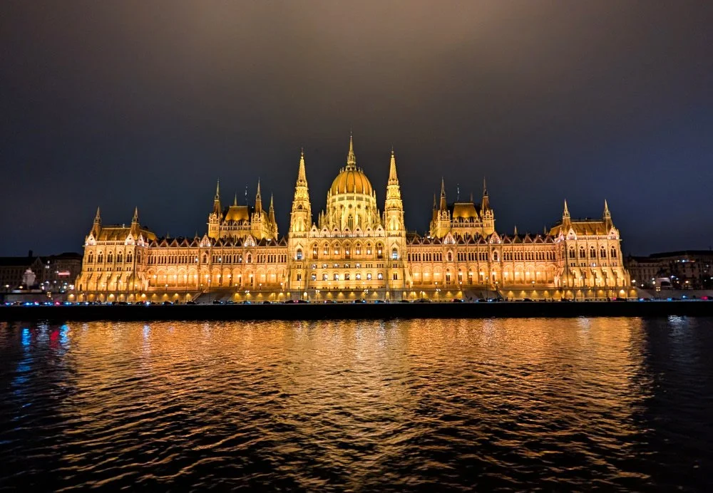 The Budapest Parliament Building as seen from the Danube at night, it glows golden and is reflected in the river water