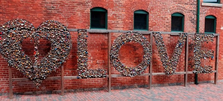 Love locks in the Distillery District. A large sculpture of a heart and the word "love" all filled with padlocks. Behind it is an old, red brick factory wall with sever al green-painted windows. The ground under it is cobblestone.