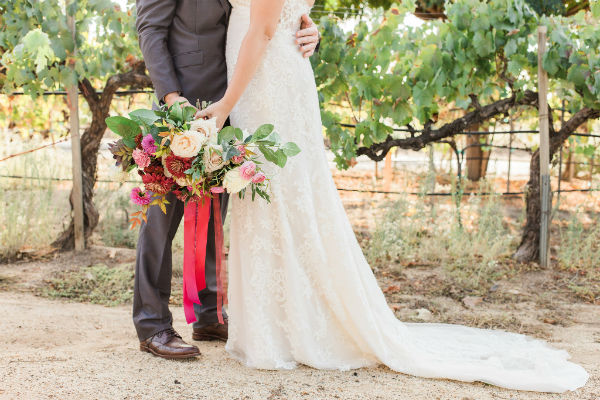 Lower half of bride and groom holding bouquet in front of vineyard row
