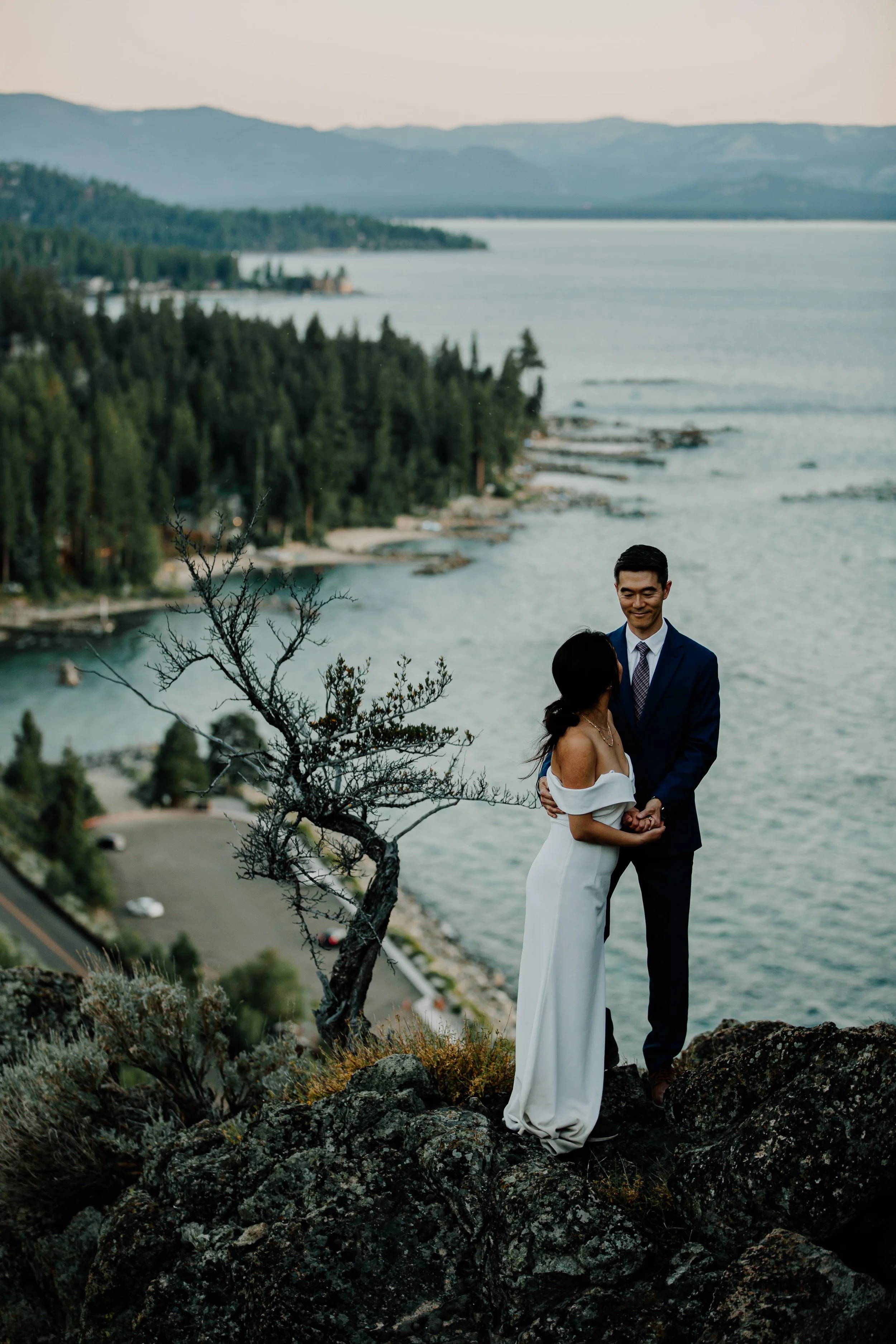 A couple dressed in formal attire standing on a rocky hilltop during sunset, with a lake and forested landscape in the background.