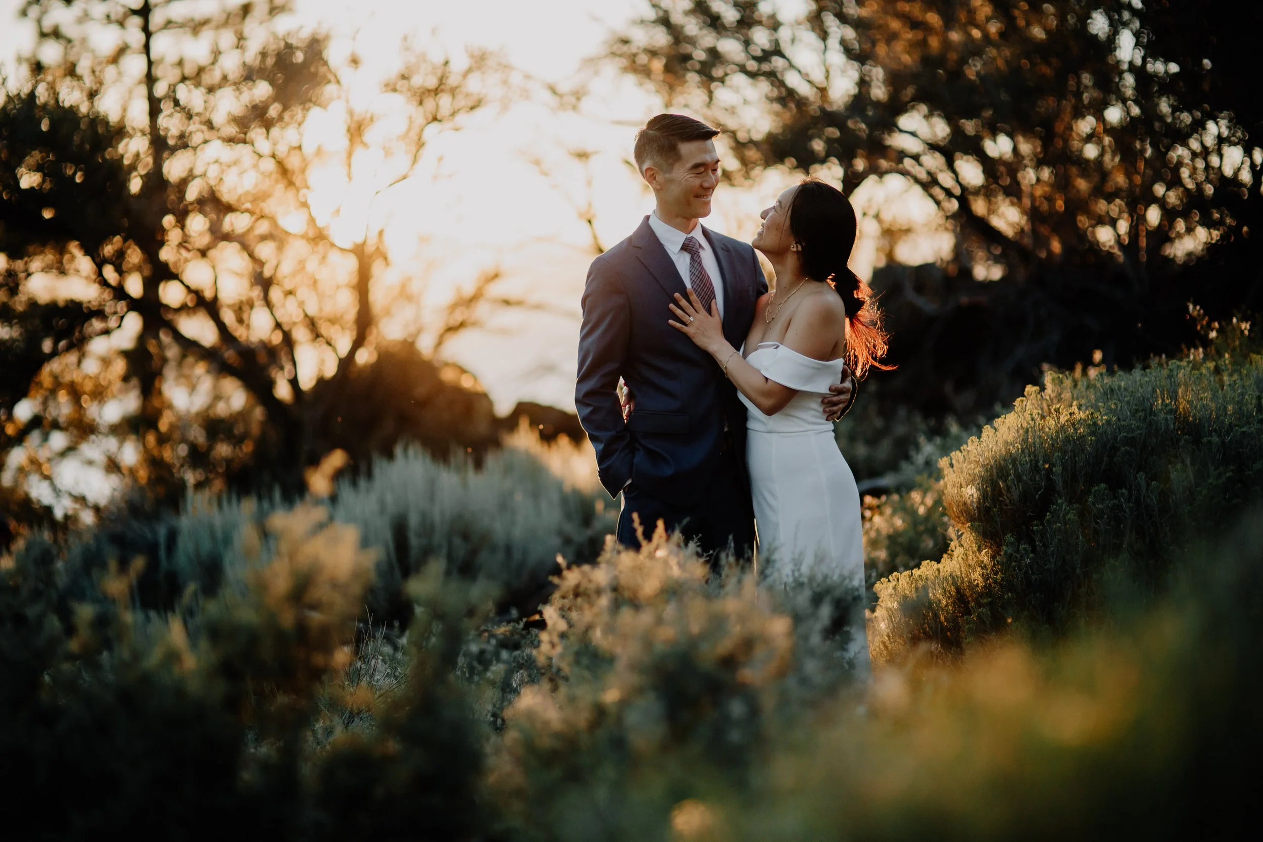 A romantic couple dressed in elegant fashion standing close together outdoors at sunset, with warm light and trees in the background.