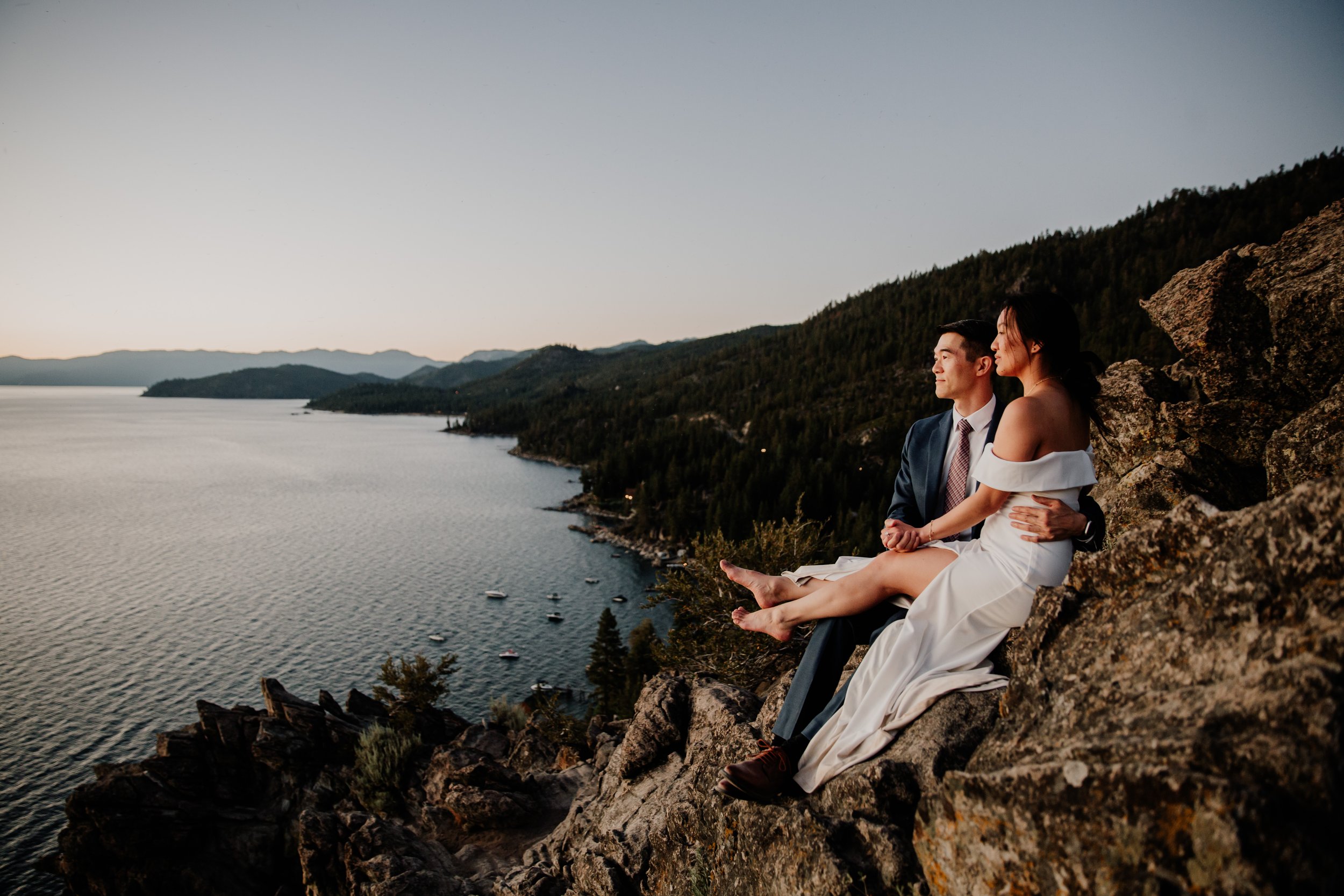 A couple in wedding attire sitting on a rocky ledge overlooking a large body of water with mountains in the distance during sunrise or sunset.