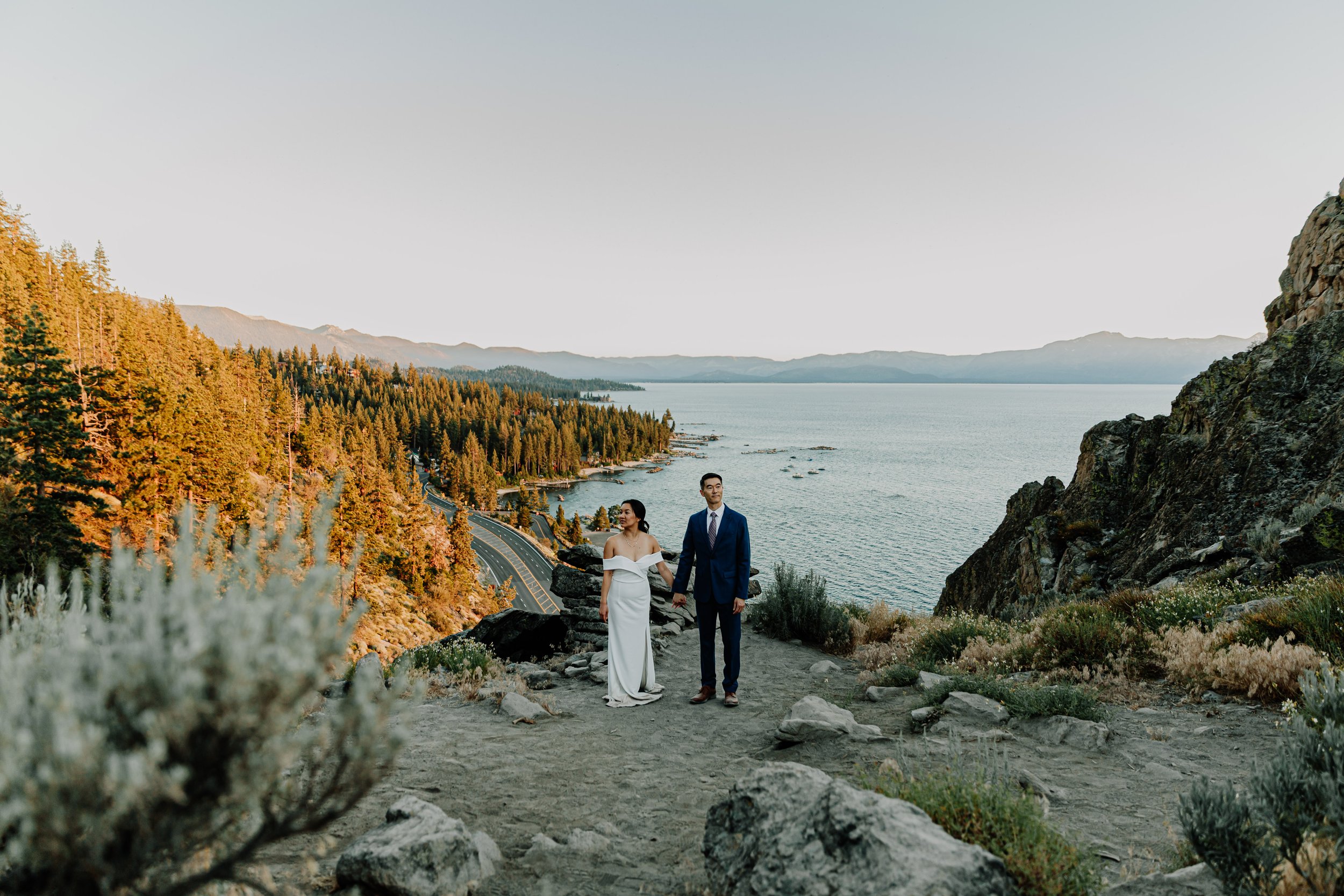 A couple stands on a rocky overlook by a lake at sunset, holding hands, with a forested hillside and mountains in the background.