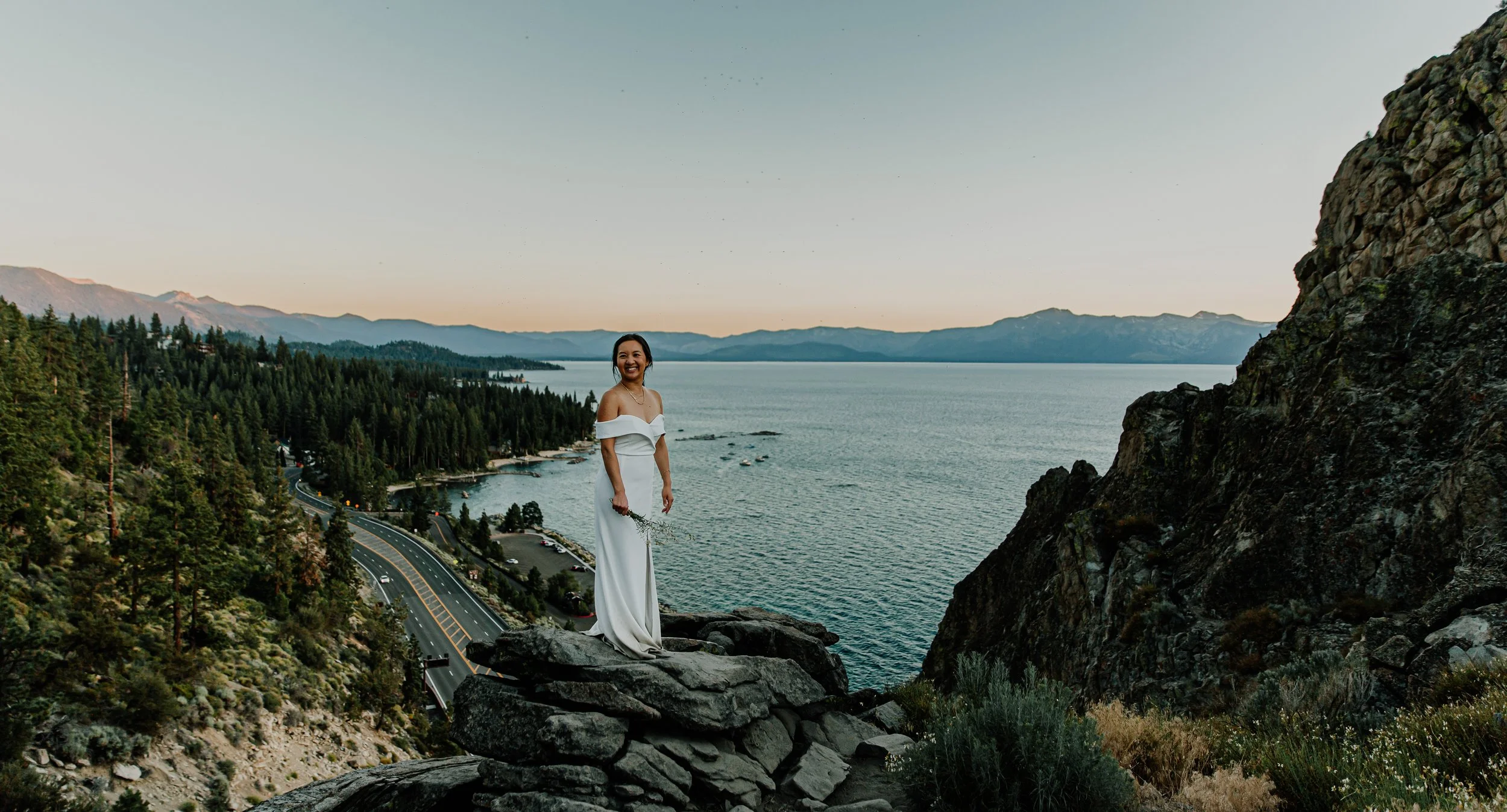A woman in a white off-shoulder dress standing on rocks by a lake with mountains and forest in the background during sunset.