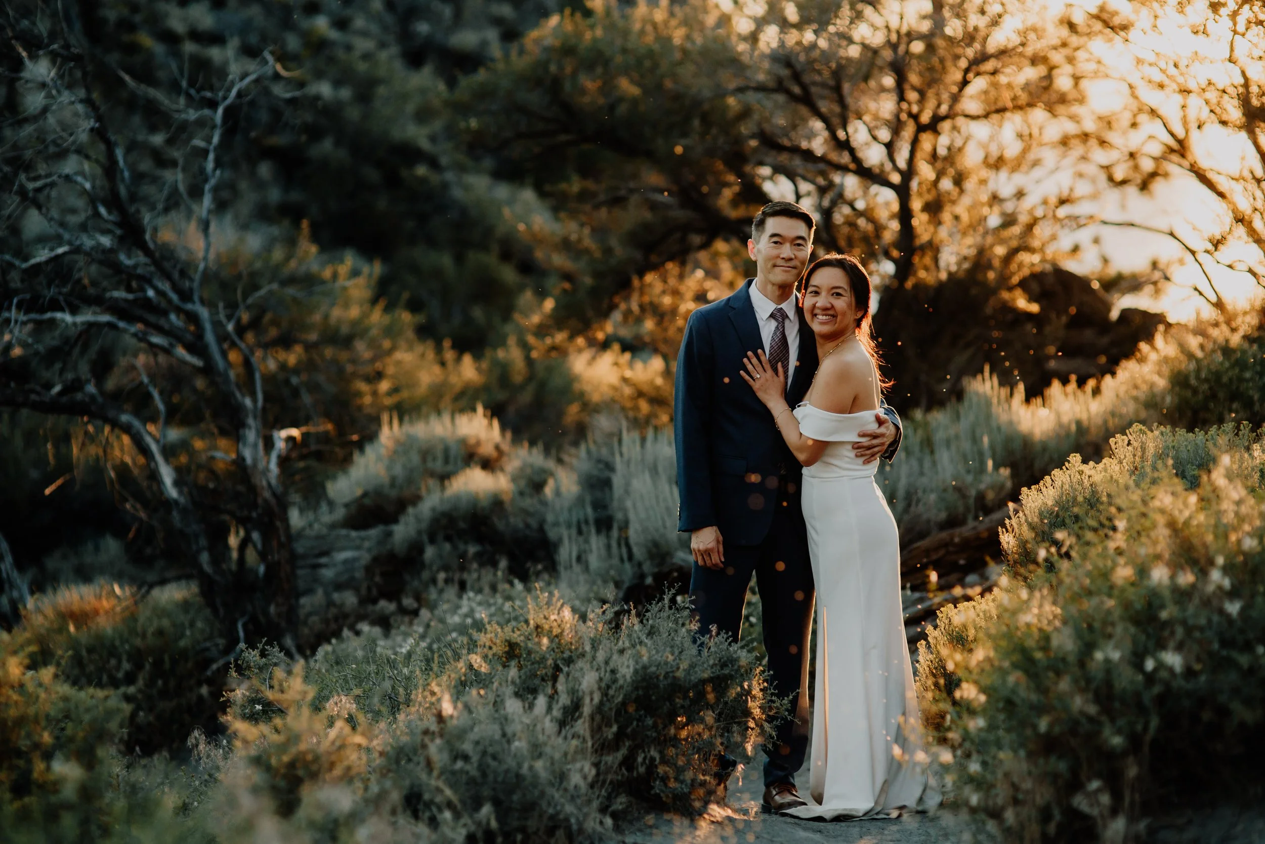 A couple standing close together outdoors during sunset, smiling and embracing, with trees and foliage in the background.