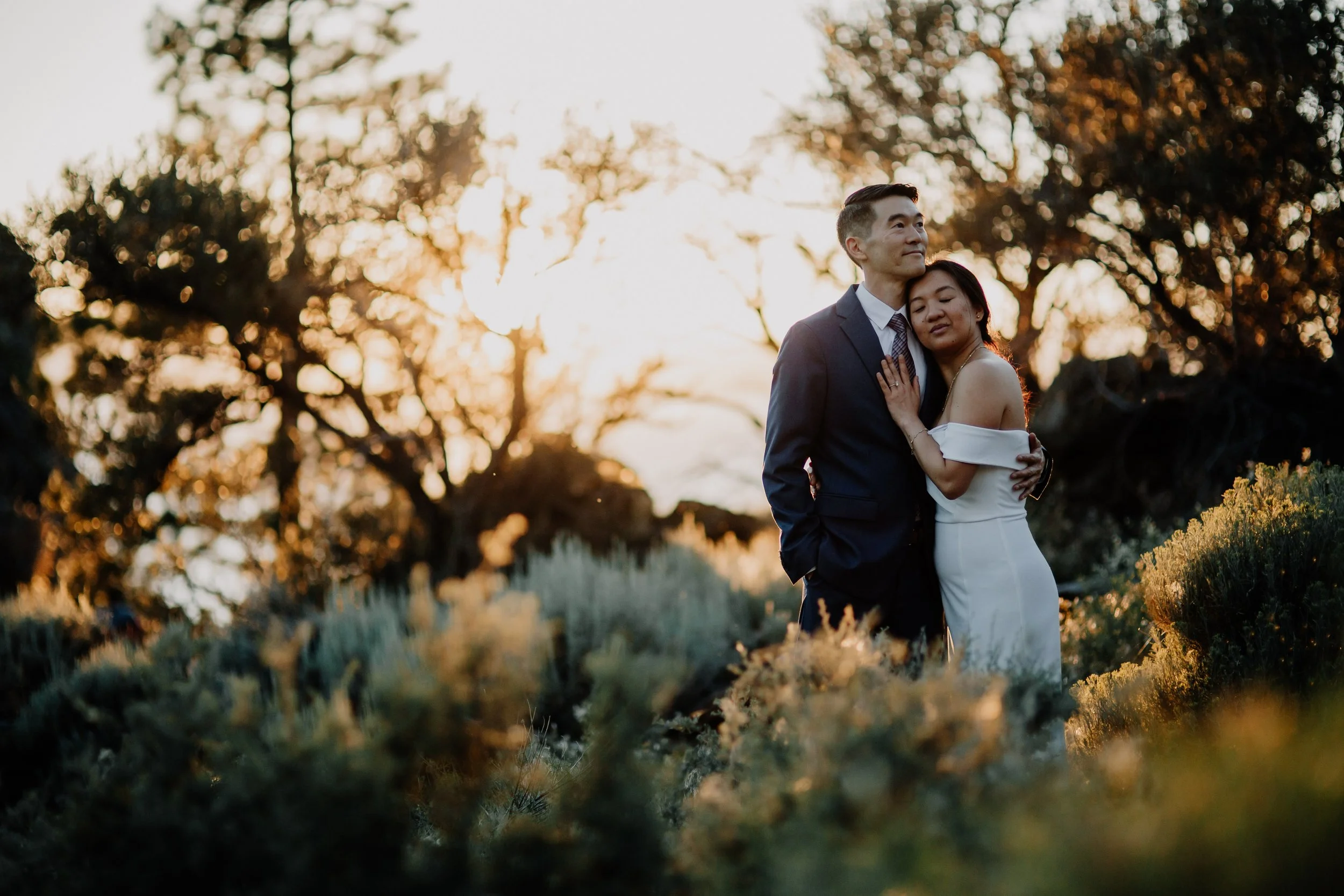 A couple dressed in formal wedding attire embracing outdoors at sunset, with trees and greenery in the background.