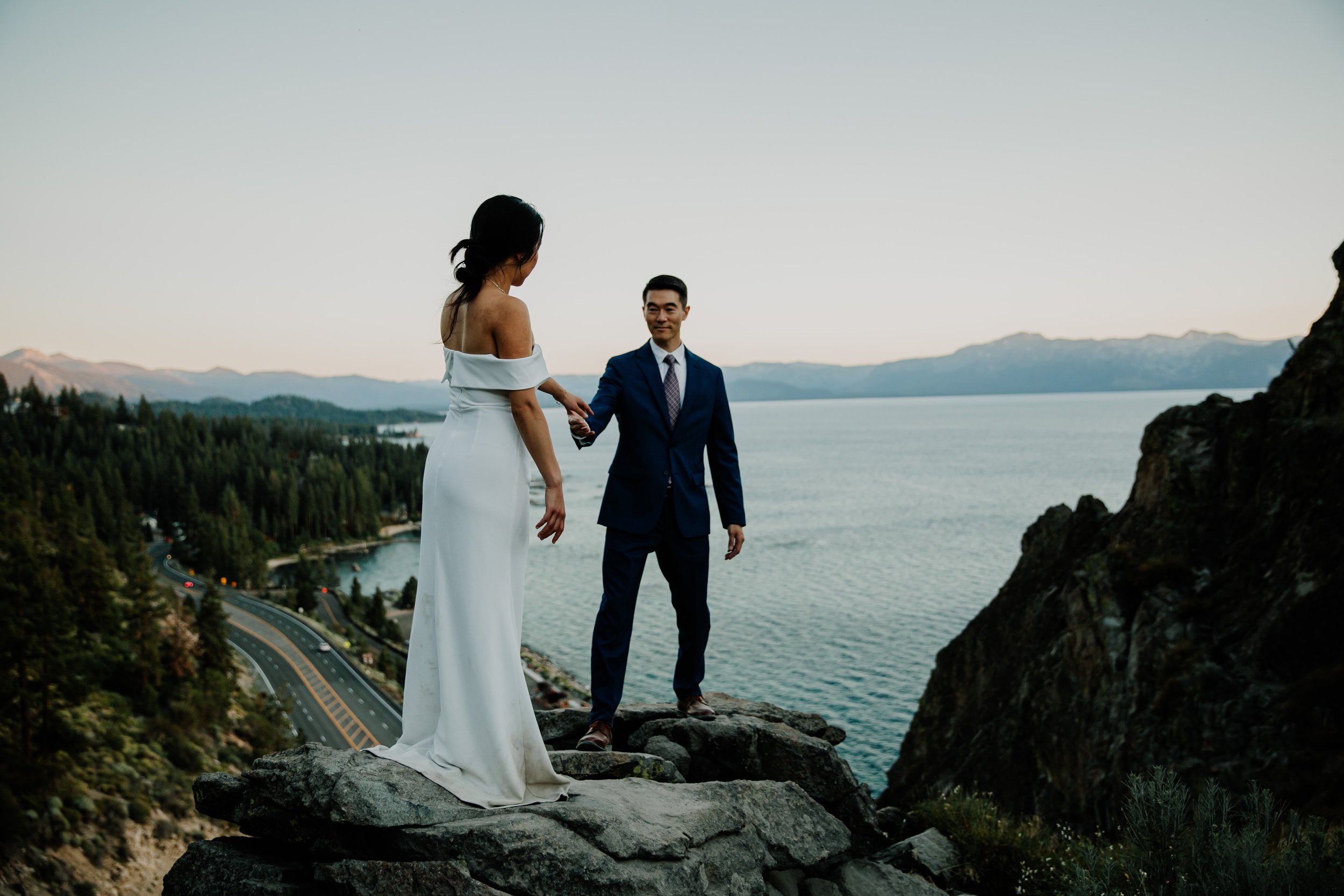 A couple in wedding attire standing on rocks with a scenic lake and mountains in the background, holding hands.