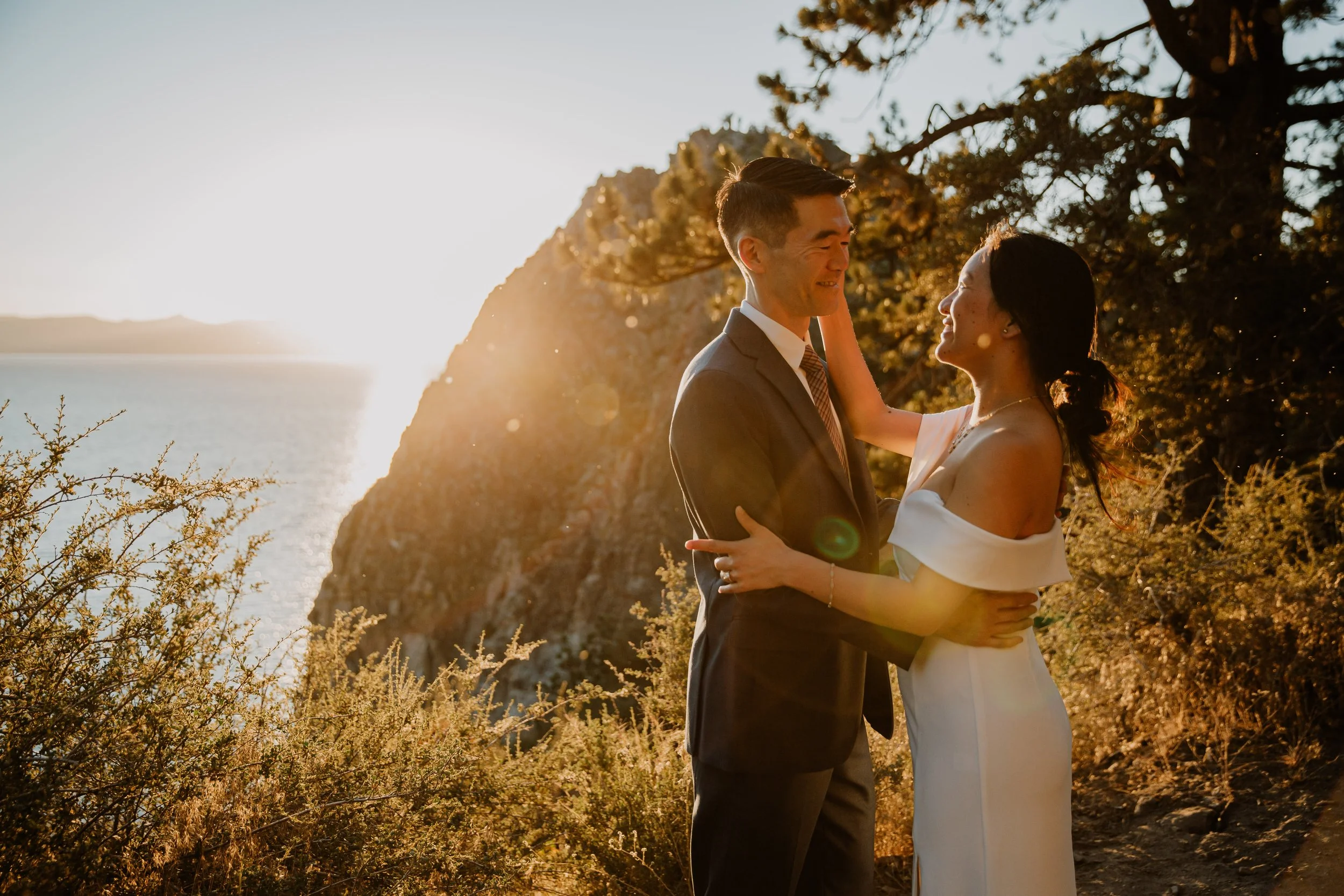 A couple dressed in formal attire embracing each other on a beach at sunset, with the ocean and rocky cliffs in the background.
