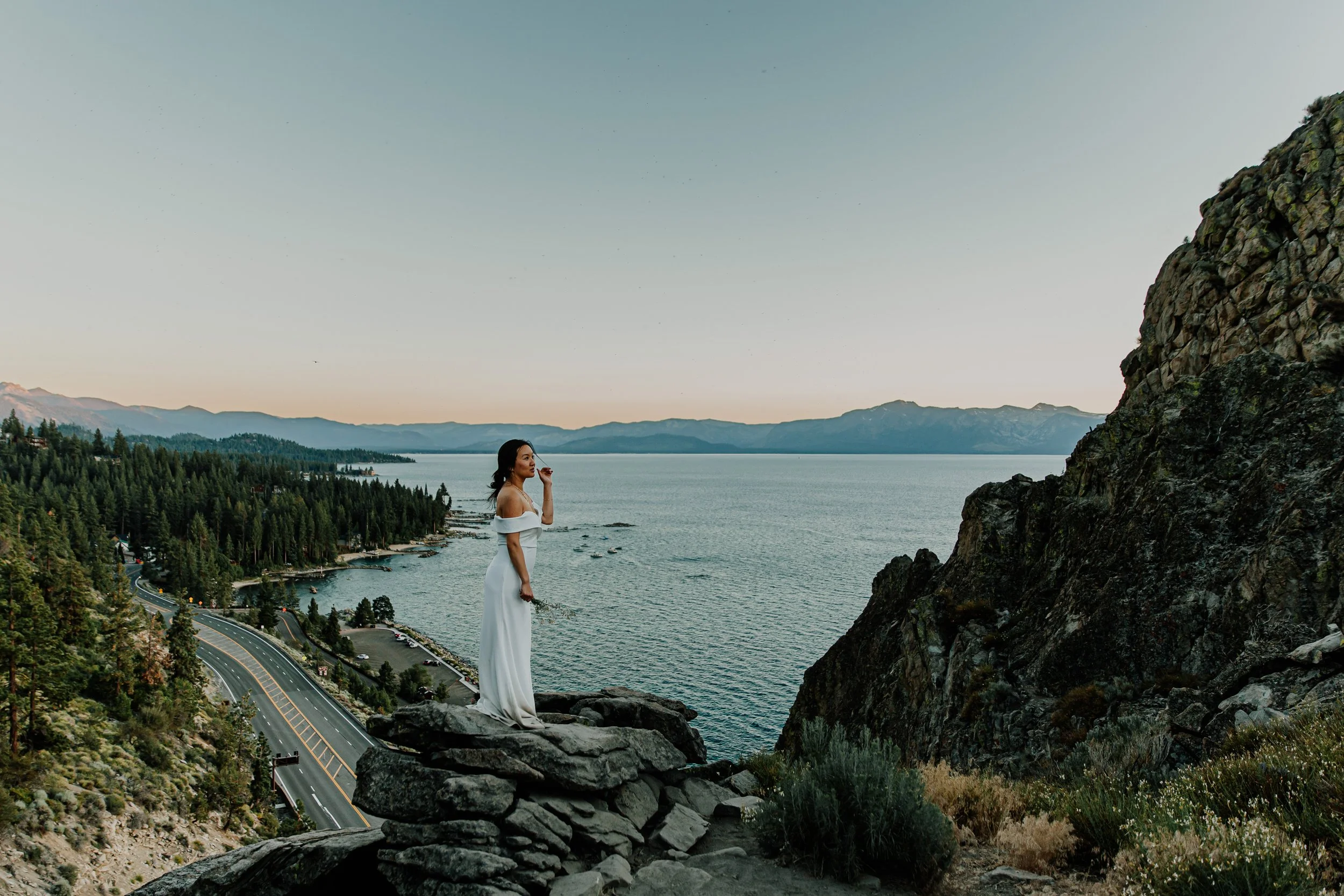 A woman in a white dress standing on rocks near a body of water with forested hills and mountains in the background.