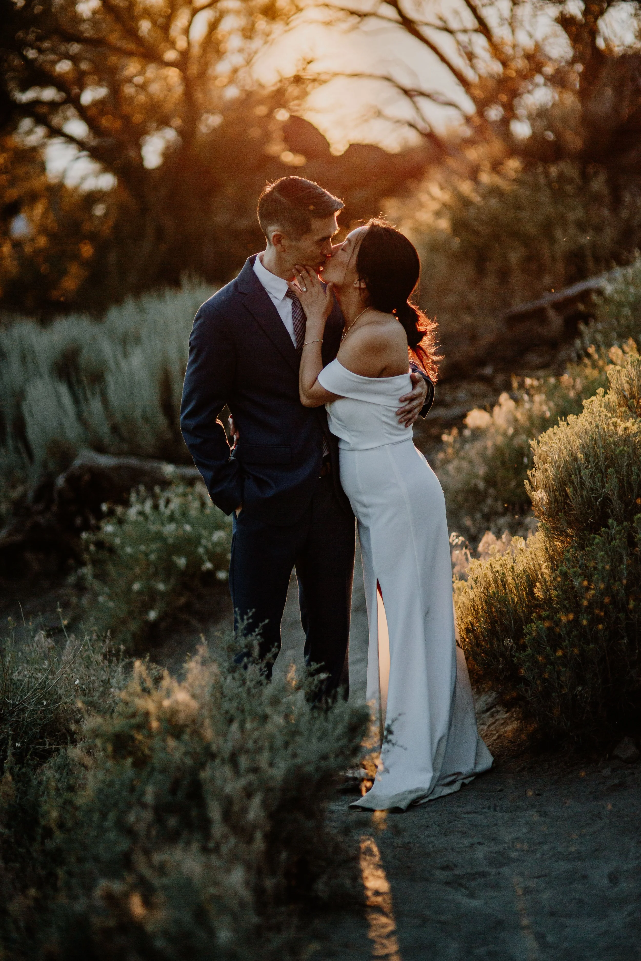 A couple in formal attire sharing a kiss outdoors during sunset, surrounded by plants and trees.