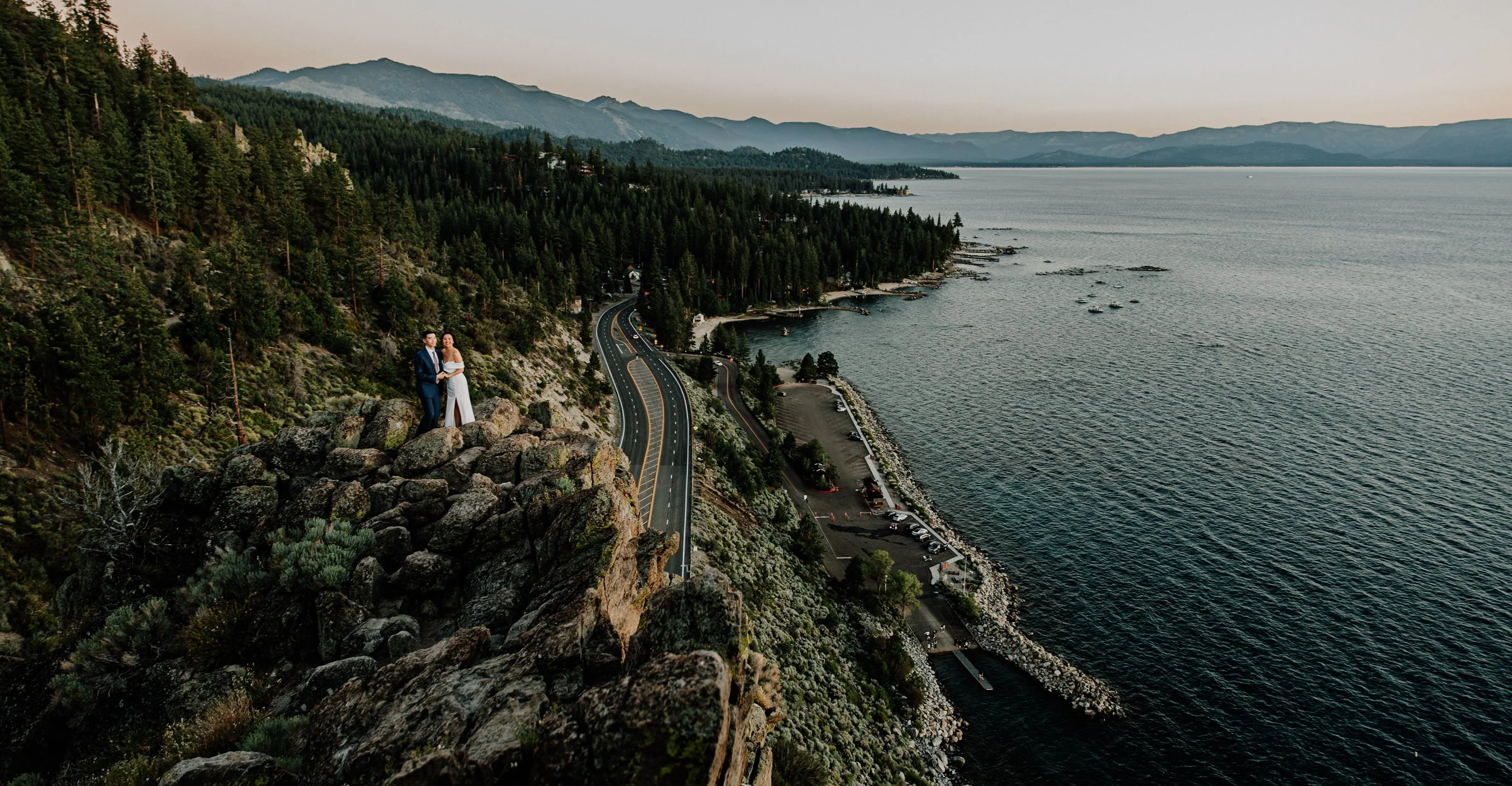 A couple dressed in formal attire standing on a rocky cliff edge overlooking a winding coastal road, forested hills, and a large body of water at sunset.