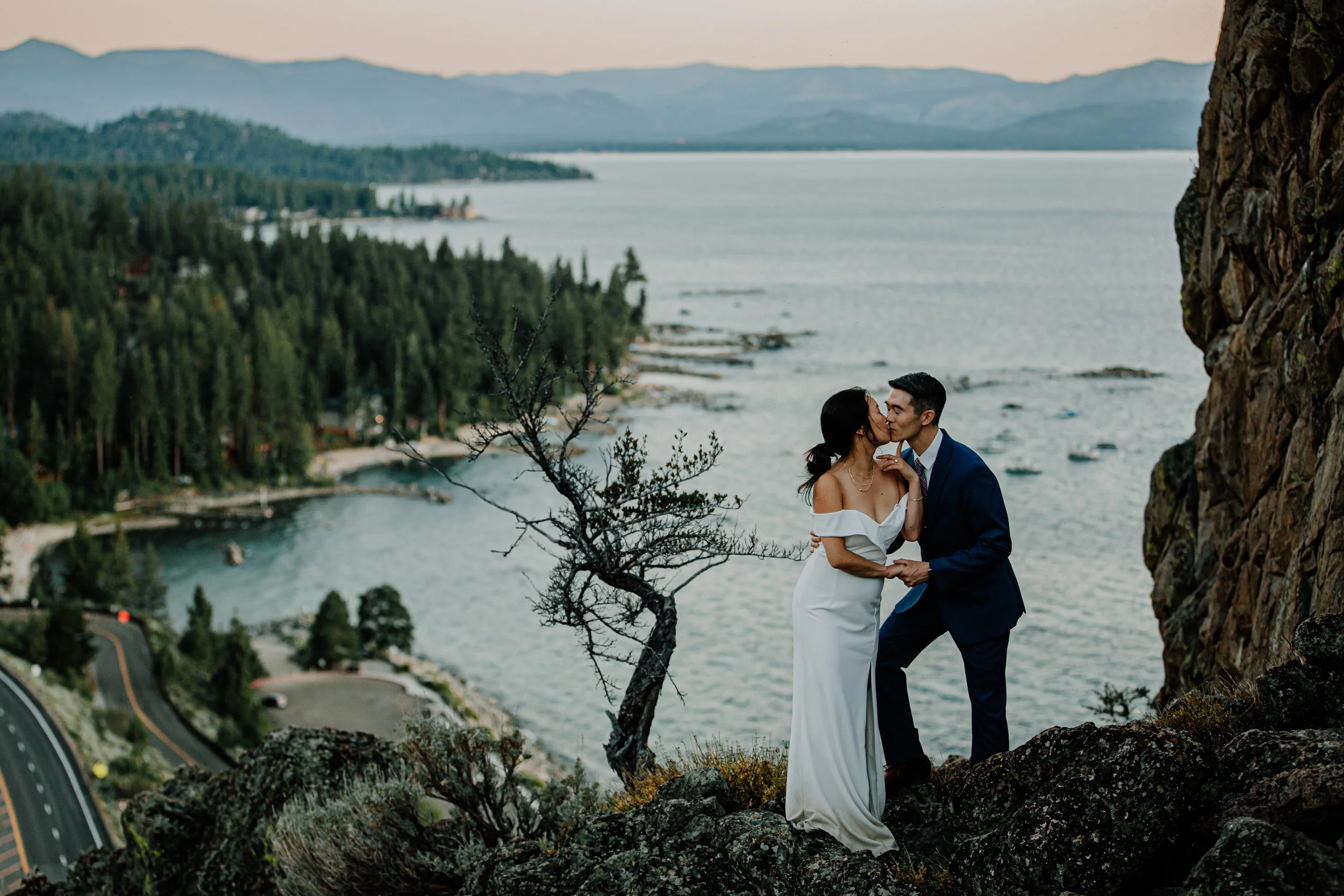 A couple dressed in wedding attire kissing on a rocky ledge overlooking a lake surrounded by trees and mountains.