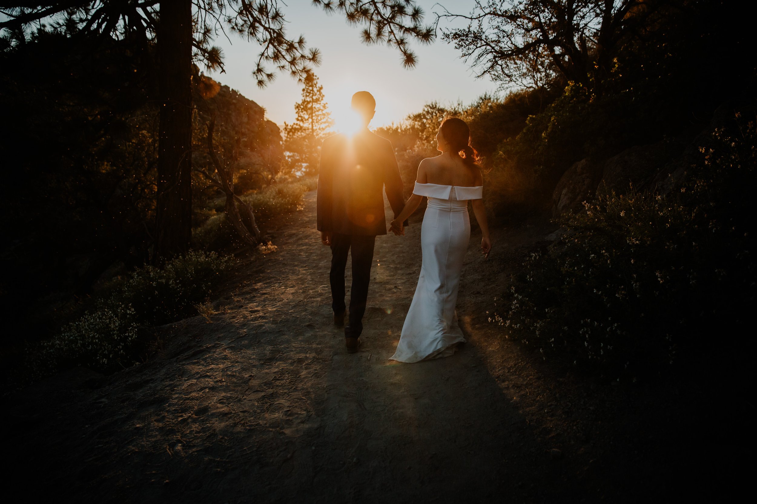 A couple in wedding attire holding hands and walking on a dirt trail in a forest during sunset.
