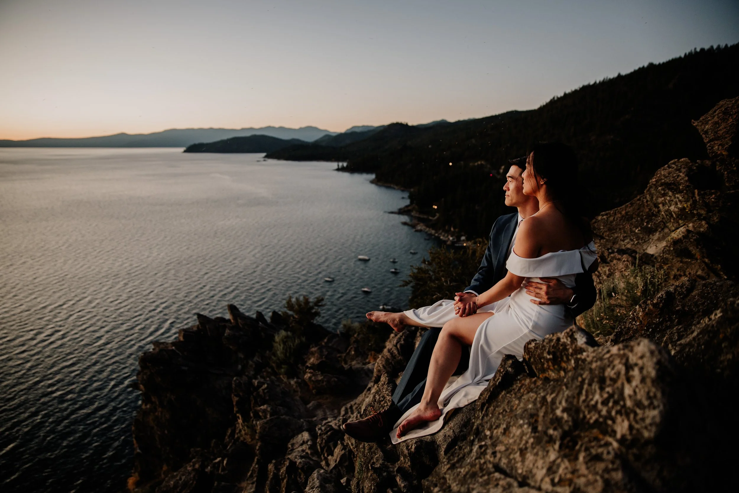 A man and woman sitting on a rocky ledge overlooking a body of water at sunset, with mountains in the distance.
