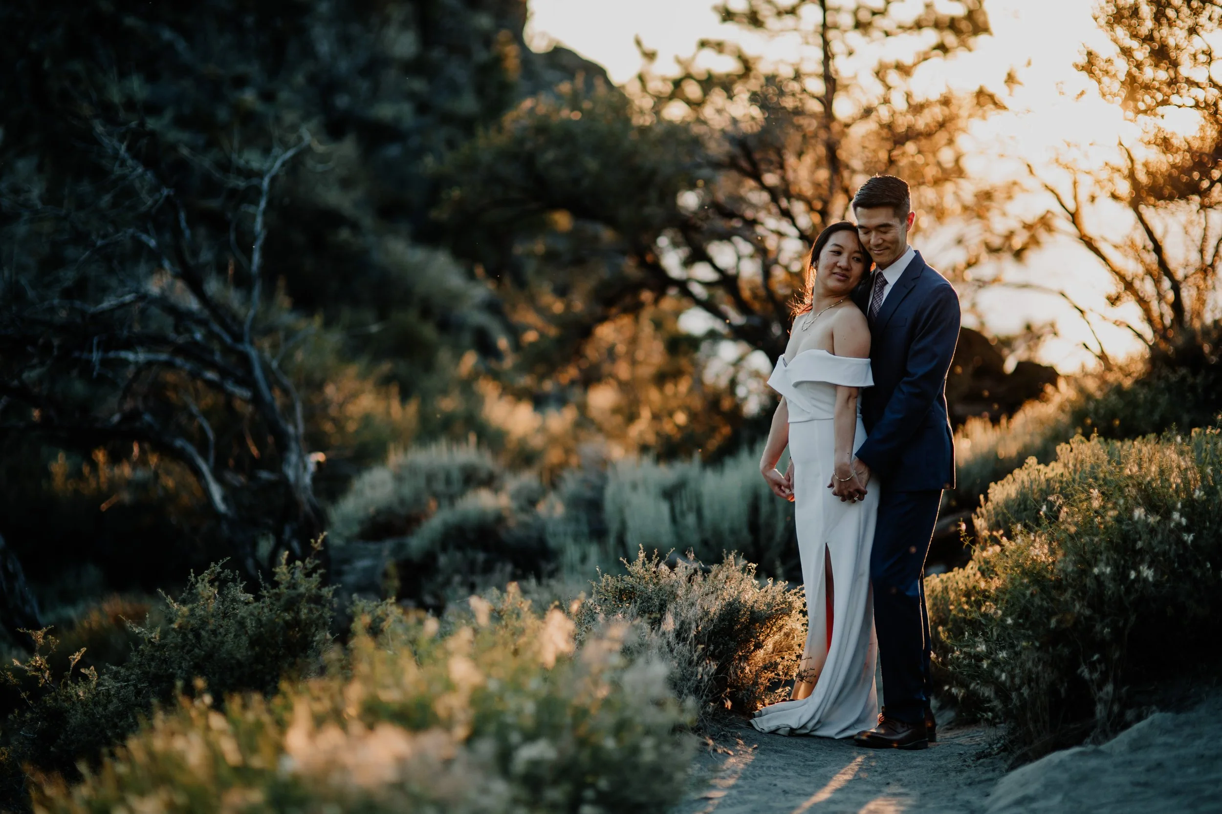 A couple in wedding attire holding hands and standing close together outdoors during sunset, surrounded by natural scenery with trees and bushes in the background.