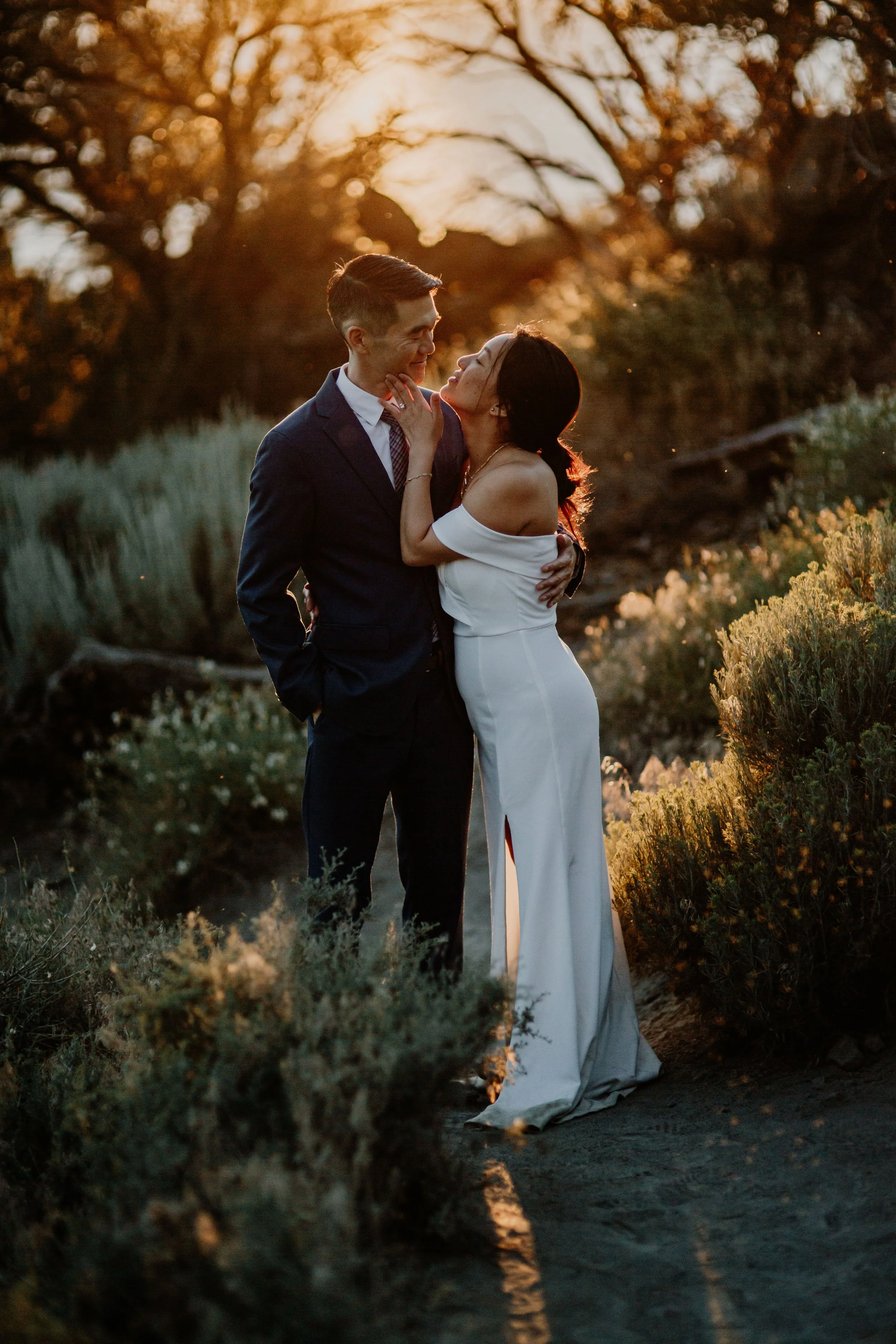 A couple dressed in formal attire standing close together outdoors at sunset, with trees and plants in the background.