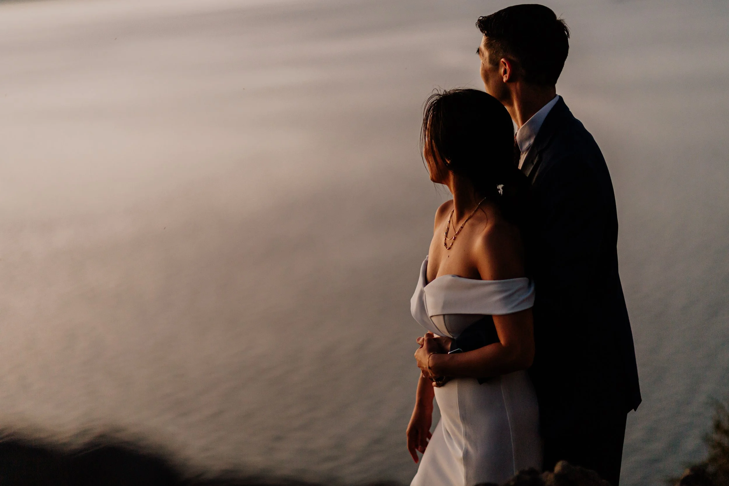 A couple standing together by the water at sunset, with the woman wearing a white off-the-shoulder dress and the man in a dark suit, embracing each other.