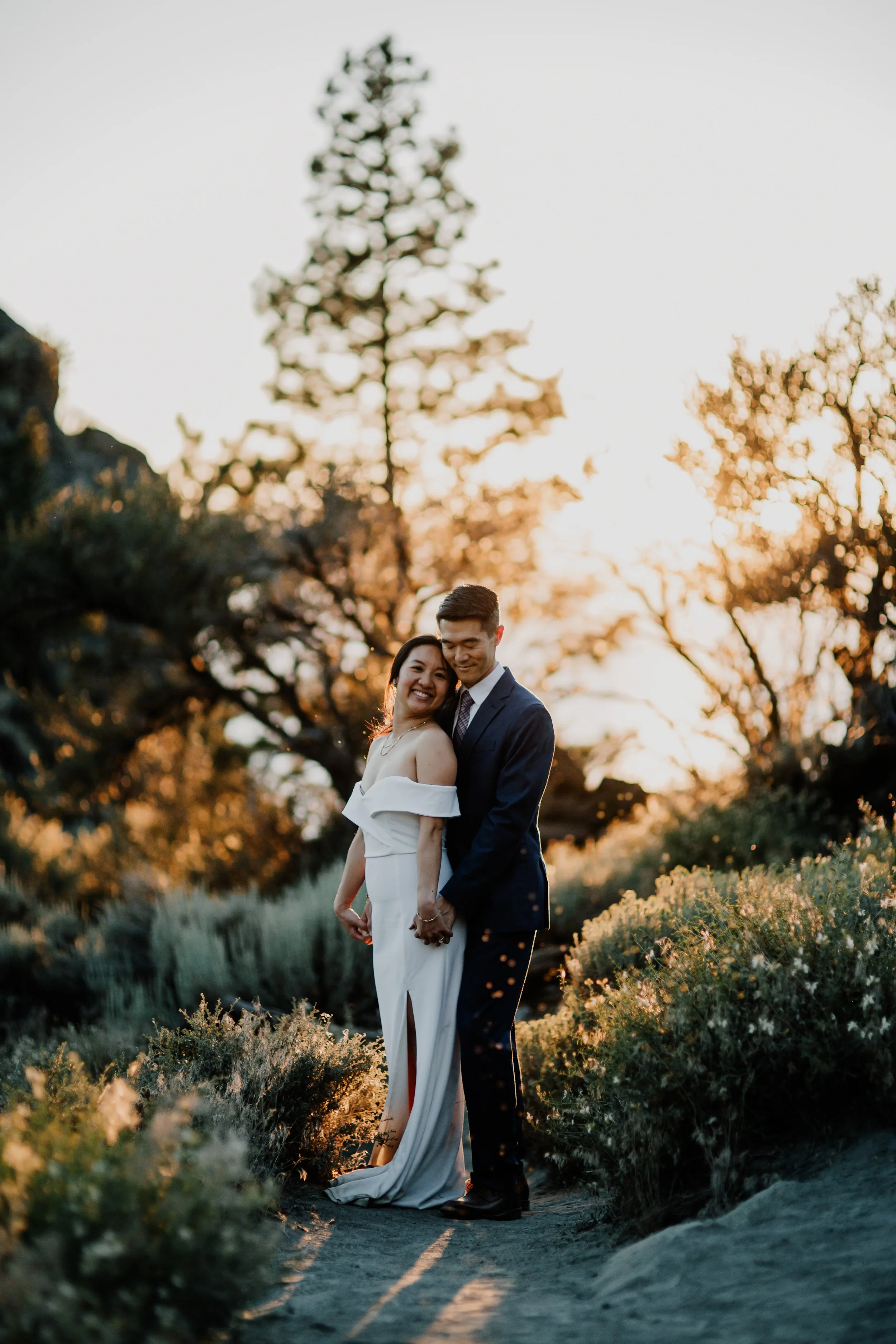 A couple dressed in wedding attire standing outdoors during sunset, holding hands, with trees and plants in the background.