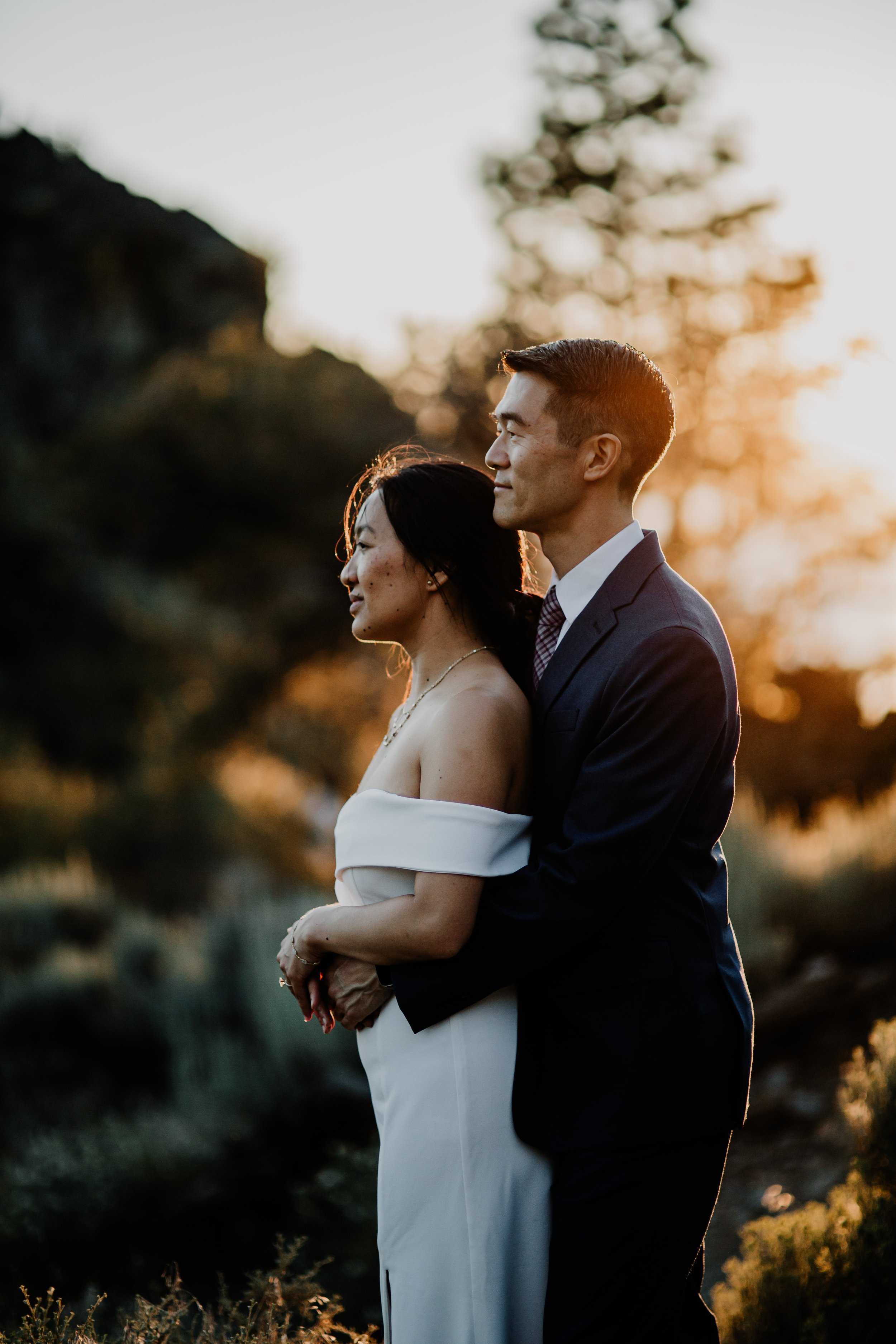 A couple dressed in wedding attire embracing outdoors during golden hour, with trees and rocks in the background.