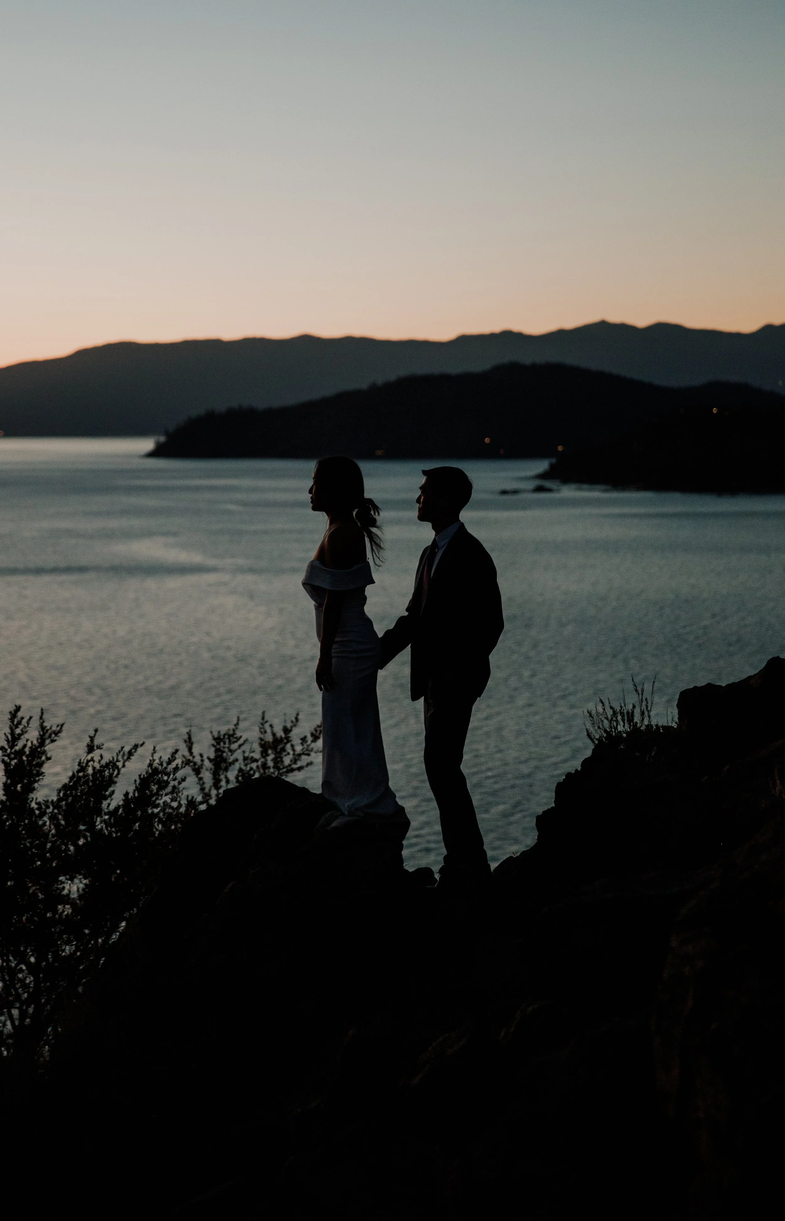Silhouette of a man in a suit and woman in an off-shoulder dress holding hands on a rocky ledge by a body of water during sunset with mountain range in the background.