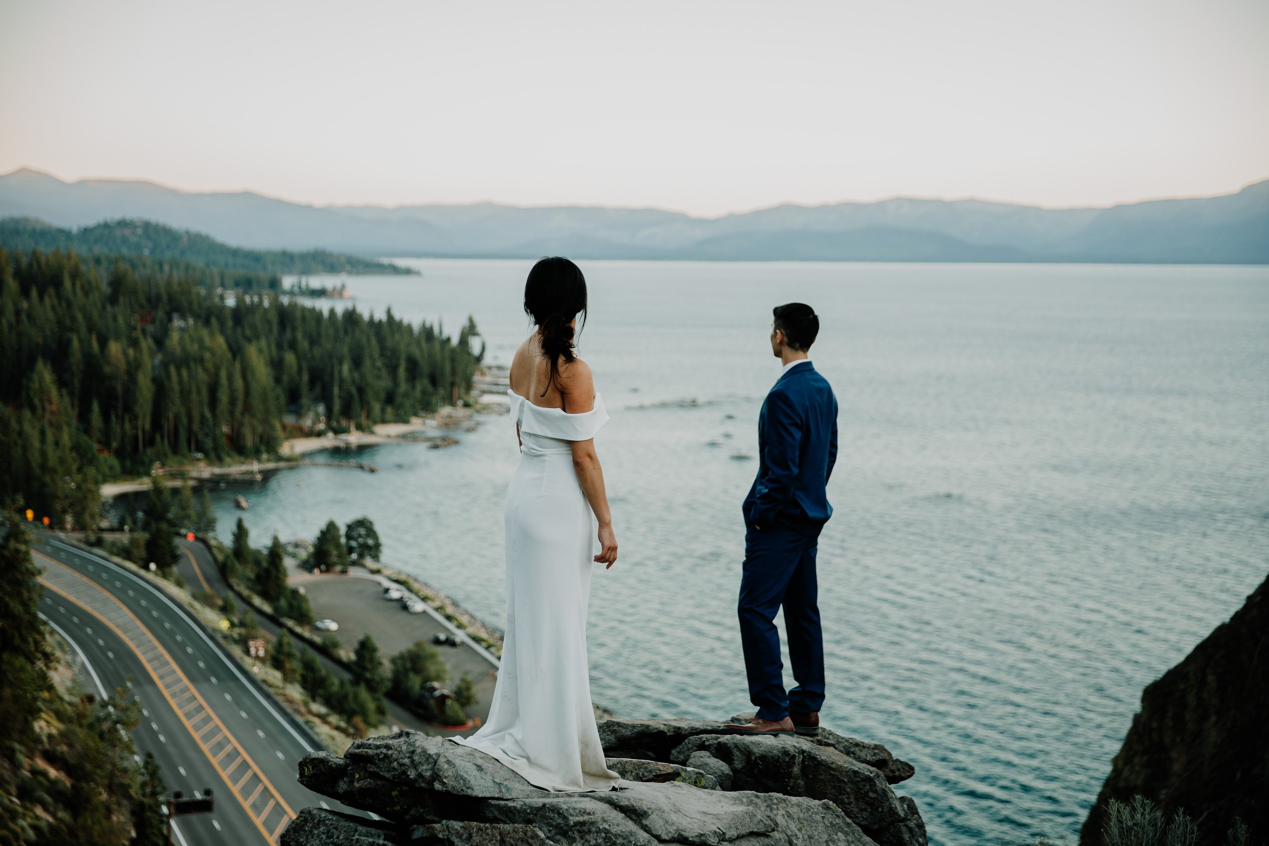 A woman in a white dress and a man in a blue suit standing on a rocky cliff overlooking a lake with mountains in the background.