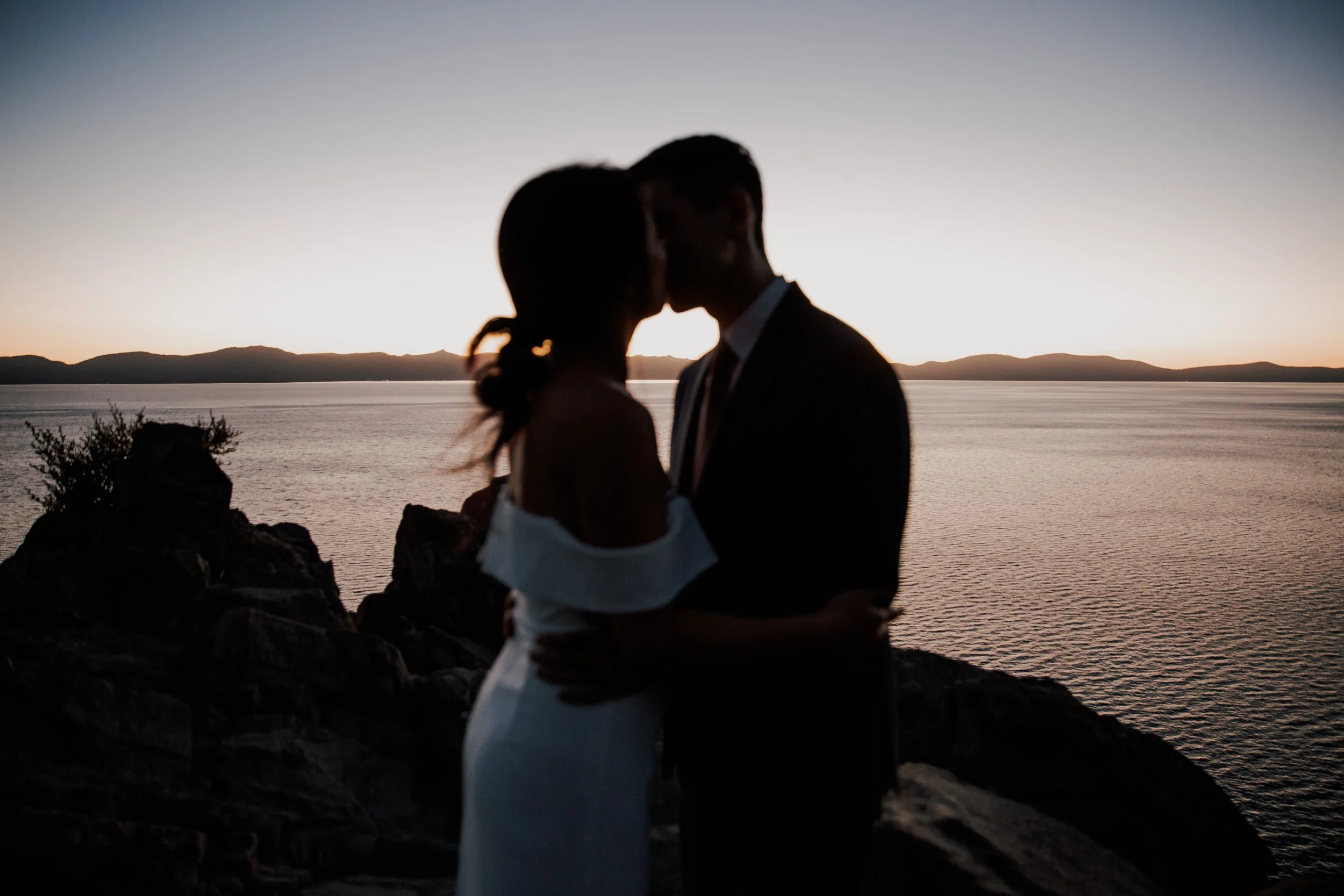 Silhouette of a couple kissing by the water at sunset with mountains in the background.