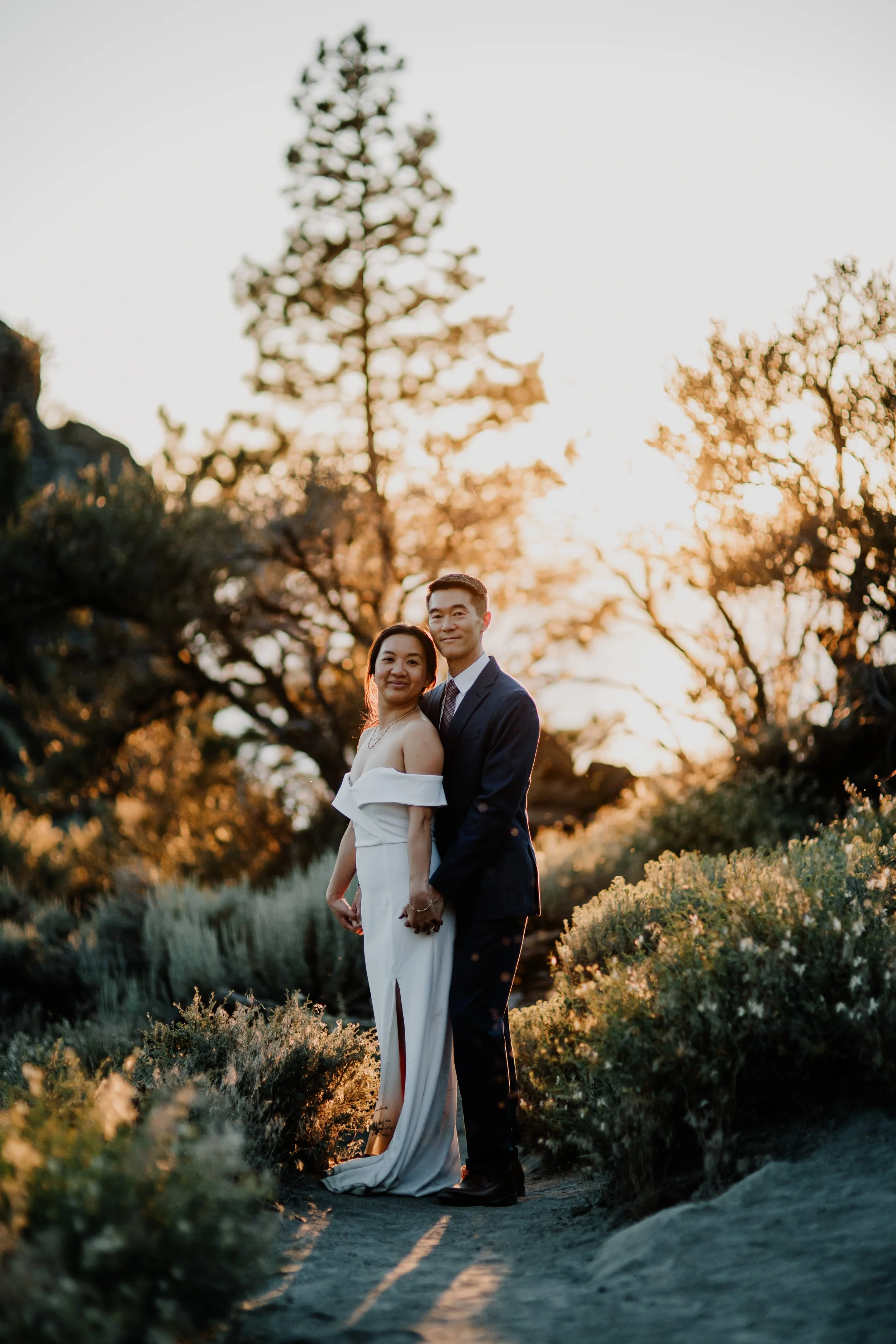 A couple dressed in formal attire stands outdoors during sunset, holding hands and smiling at the camera.