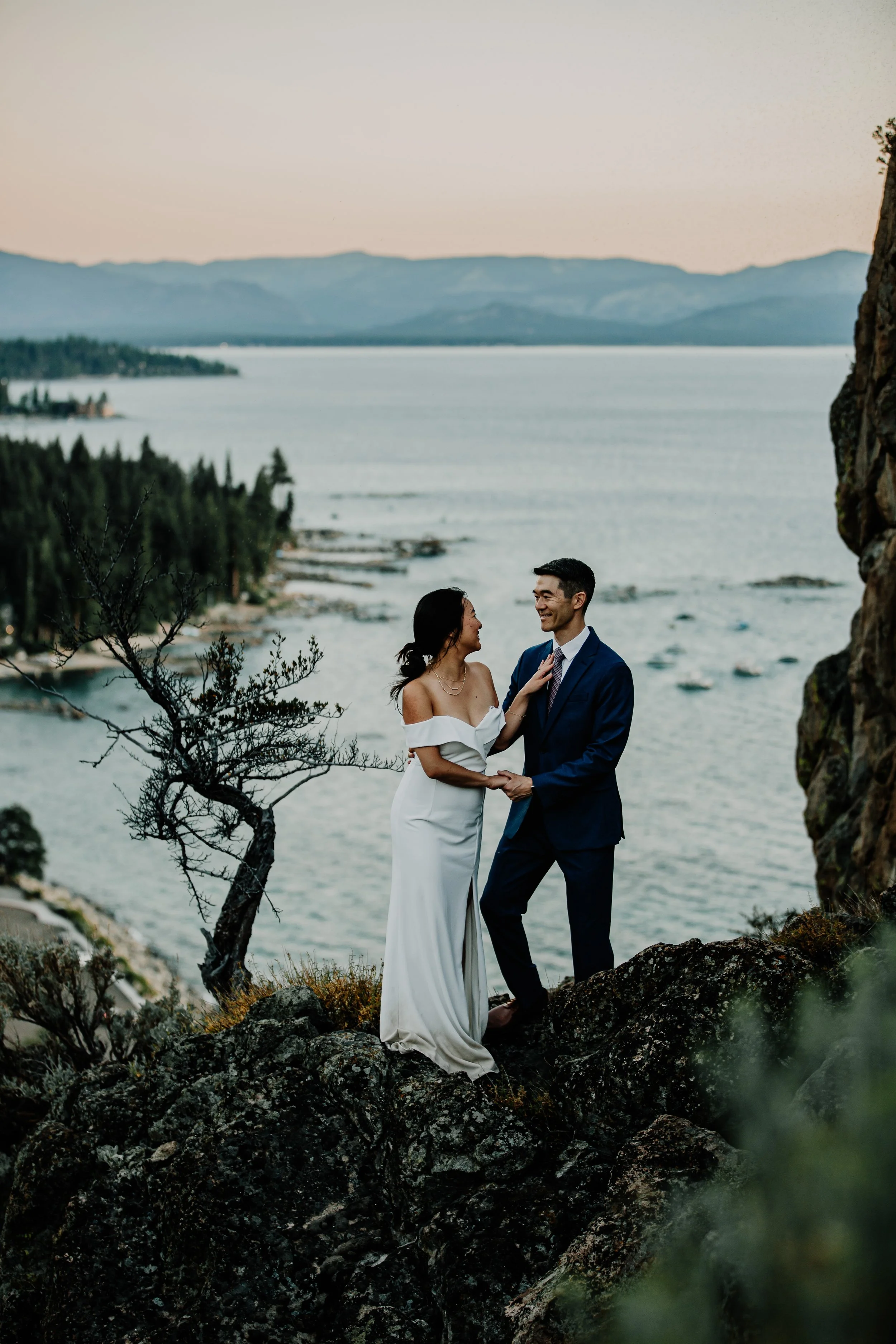 A couple dressed in wedding attire standing on rocks by a lake with mountains in the background, during sunset.