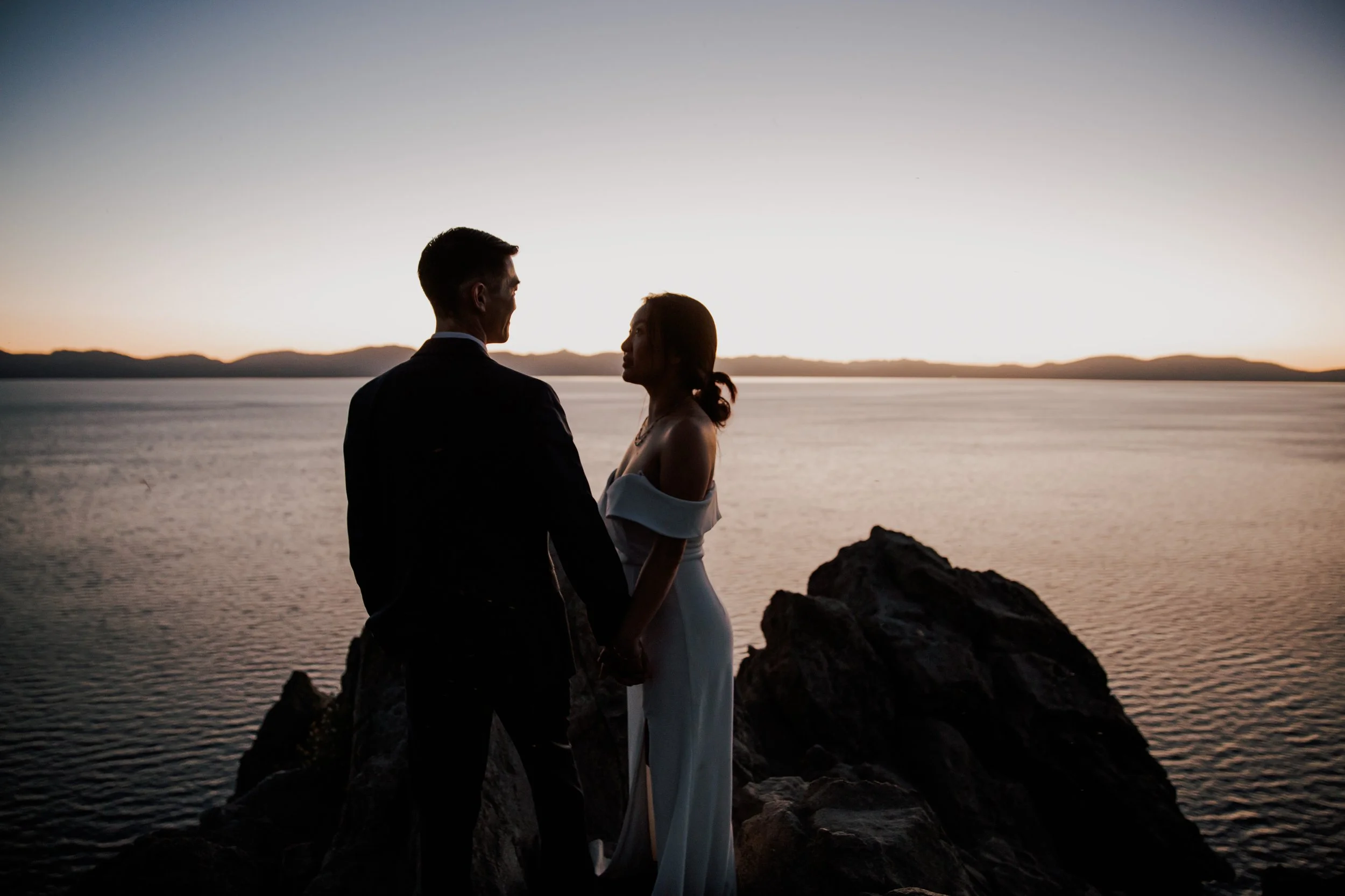 A couple holding hands at sunset by the water on rocks, with a mountain range in the background.