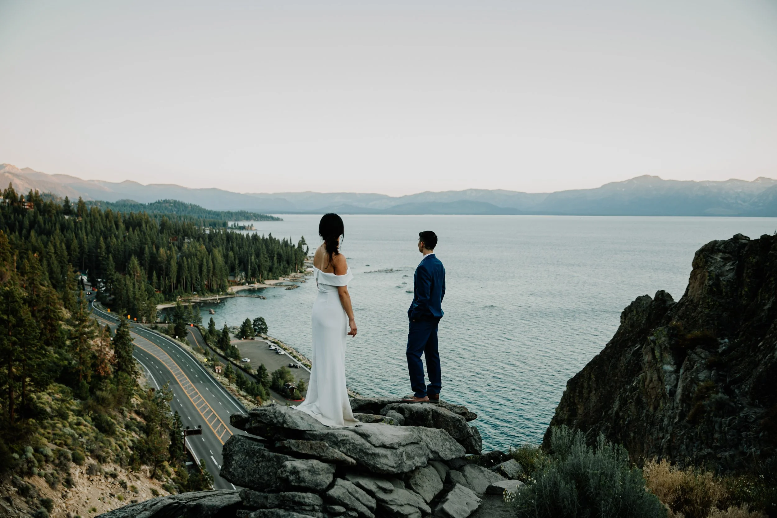 A bride and groom standing on a rocky cliff overlooking a lake and forested landscape.