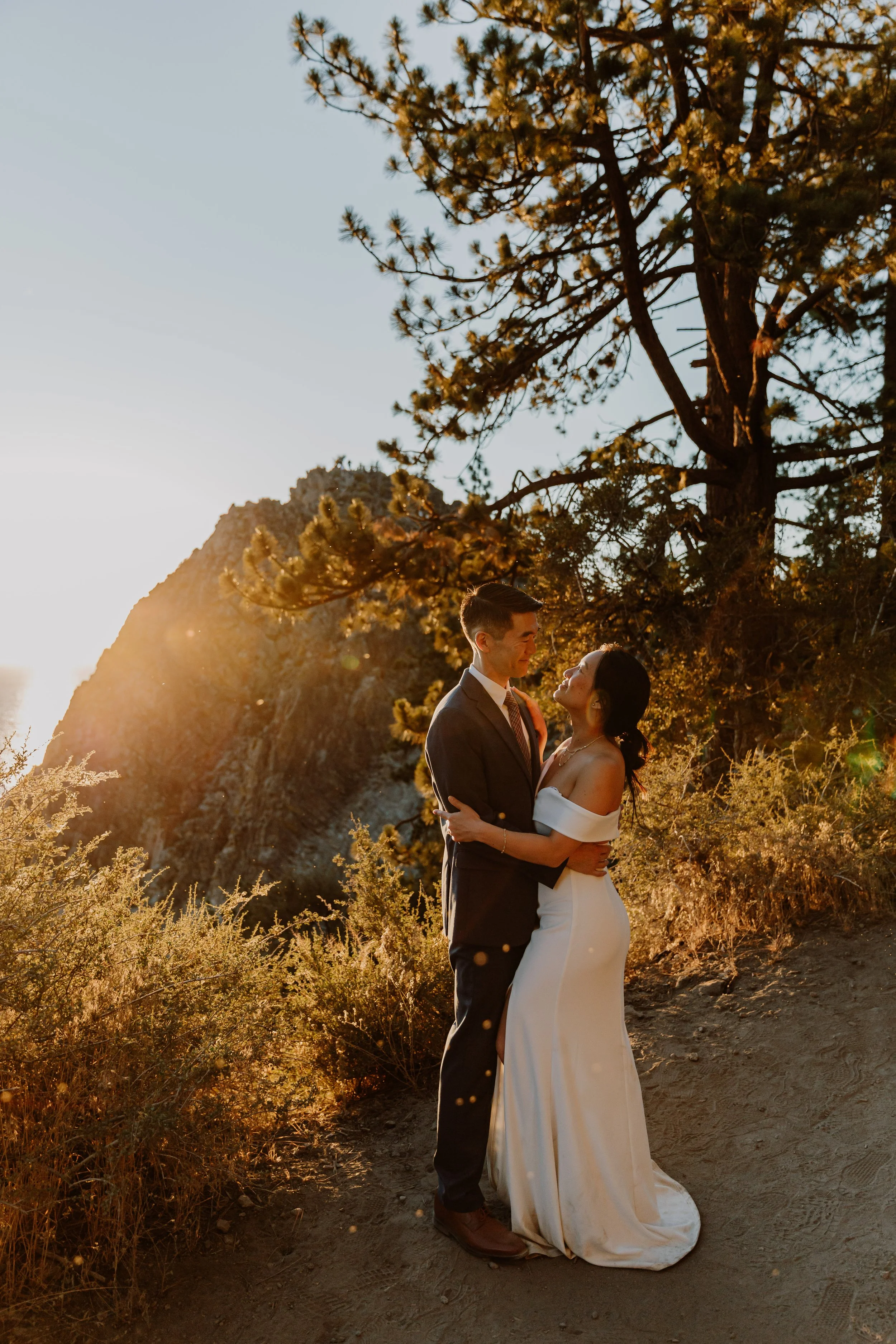 A couple in wedding attire standing on a dirt path near a pine tree, embracing and smiling at each other during sunset with mountains and the ocean in the background.