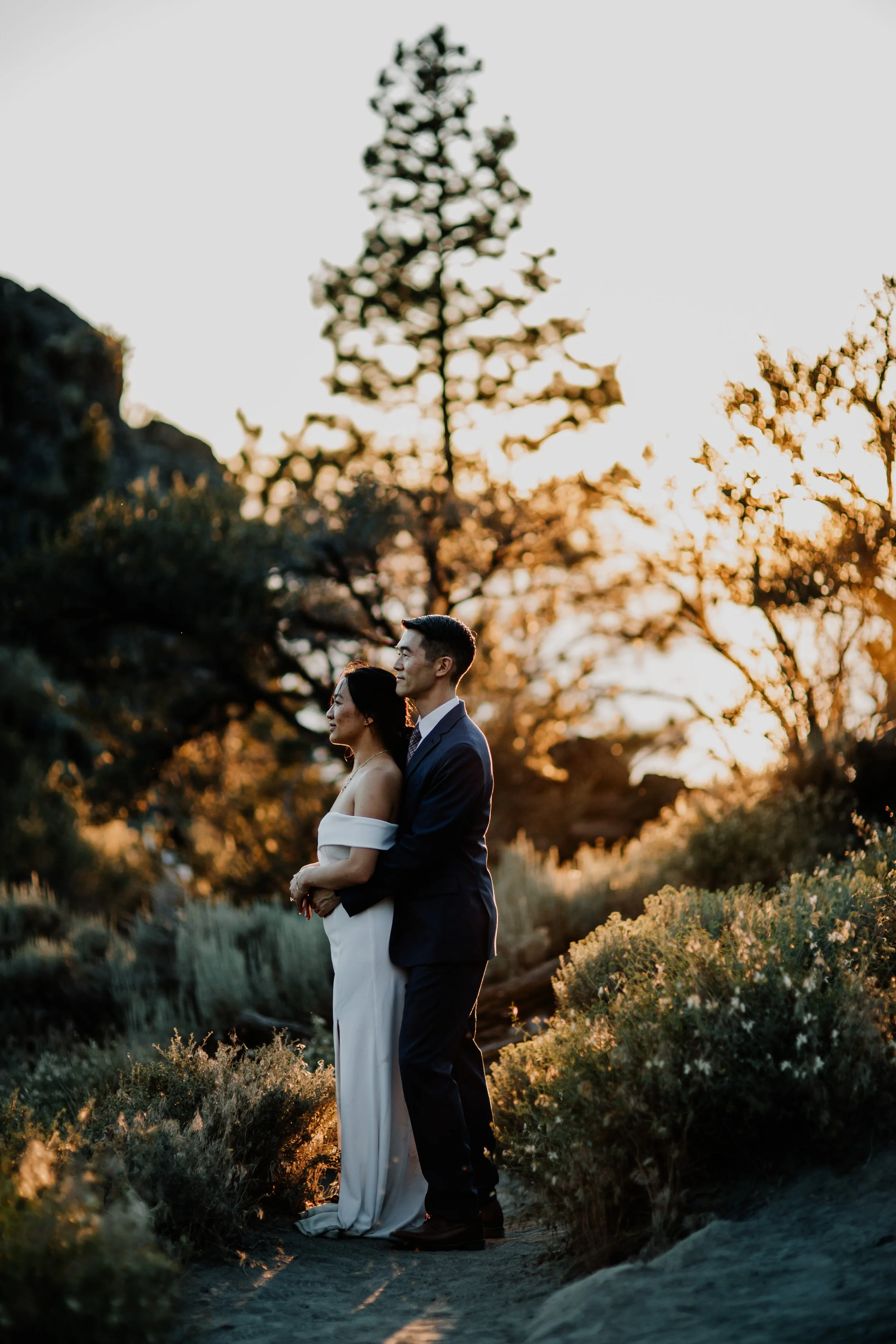 A couple in wedding attire stands outdoors during sunset, with trees and bushes in the background, embracing and gazing into the distance.
