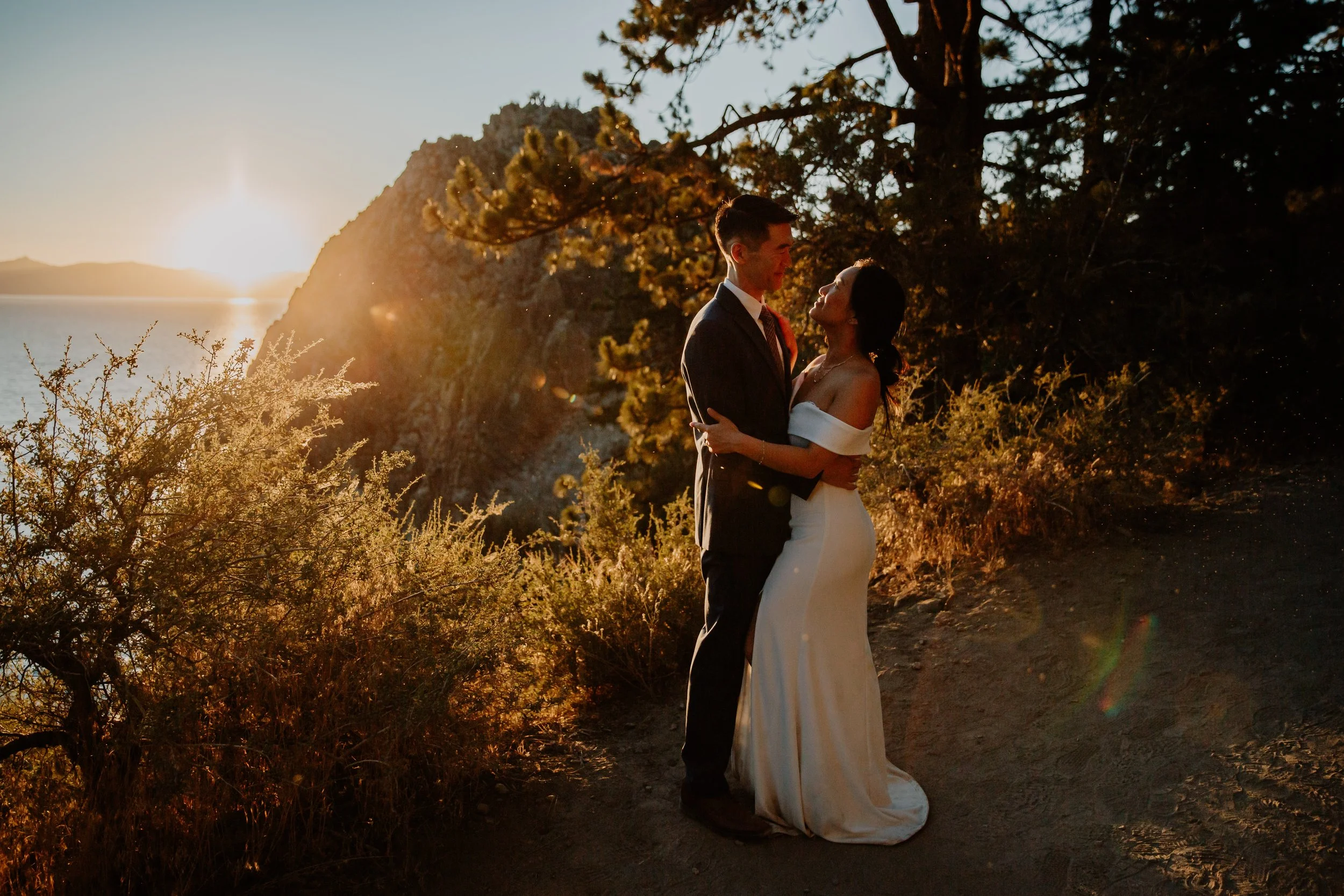 A couple in wedding attire standing close together outdoors during sunset, embracing with nature and a body of water in the background.