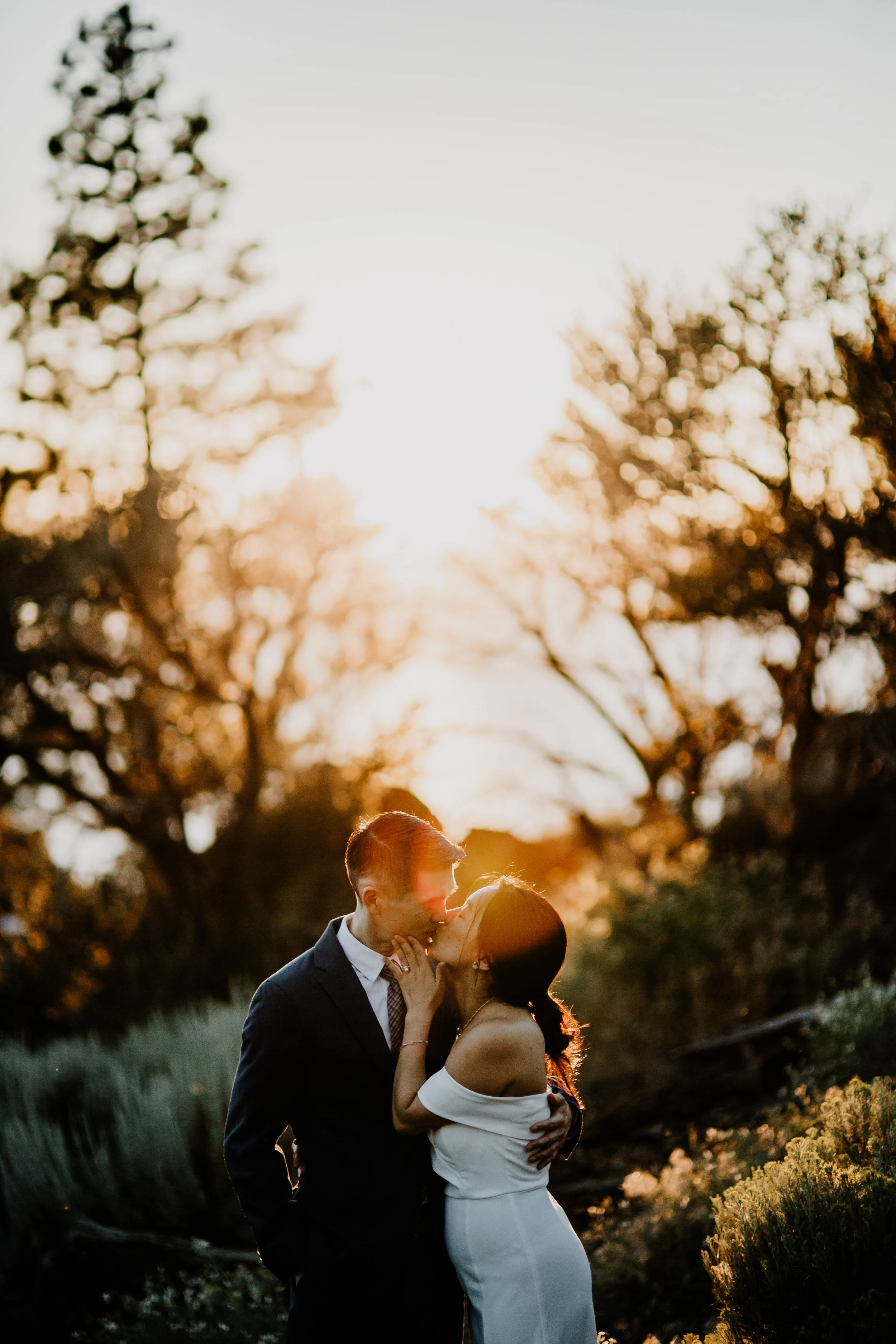 A couple in wedding attire sharing a kiss outdoors during sunset with trees in the background.
